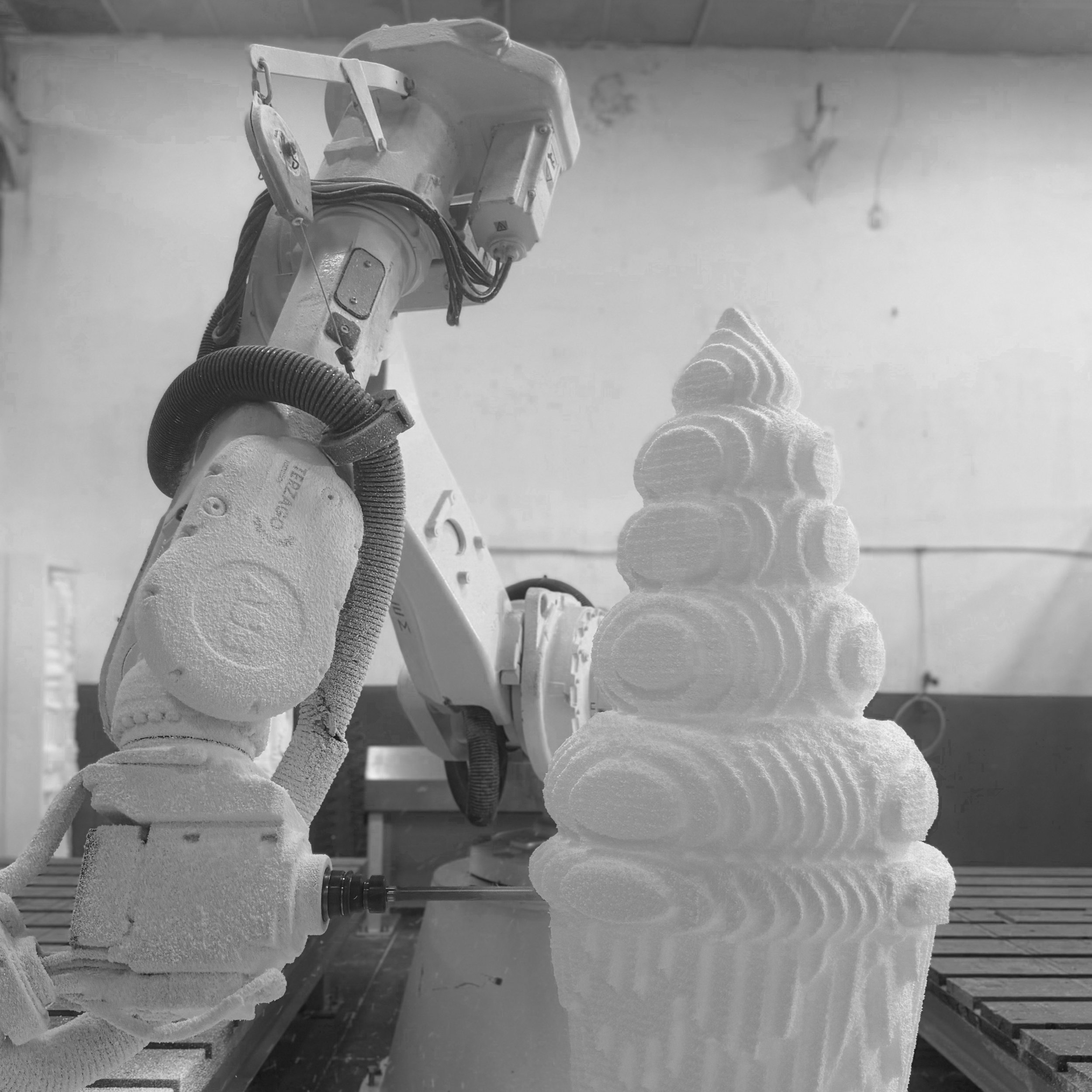 A robotic arm with a textured surface operating in a laboratory or workshop setting. The arm has a chain of cables connected to its body and is engaged in an activity alongside foam or soft material objects, with shelving and tools faintly visible in