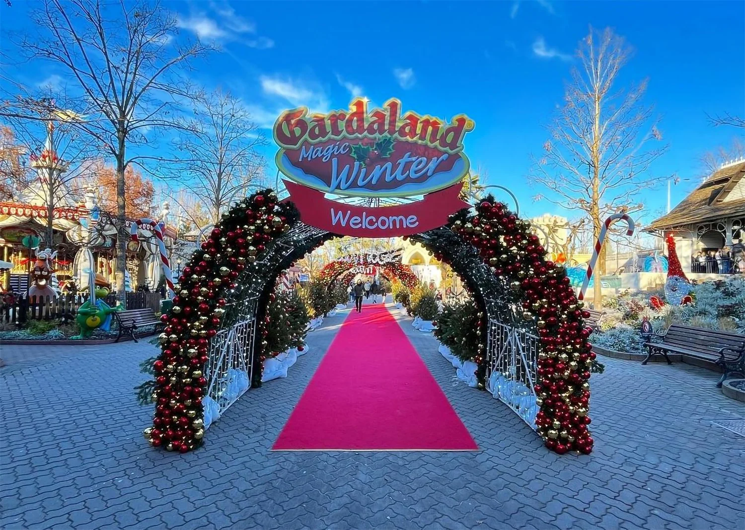 Entrance decorated with Christmas ornaments and a red carpet, leading into Gardaland Magic Winter theme park during the daytime, with festive decor and clear blue sky.