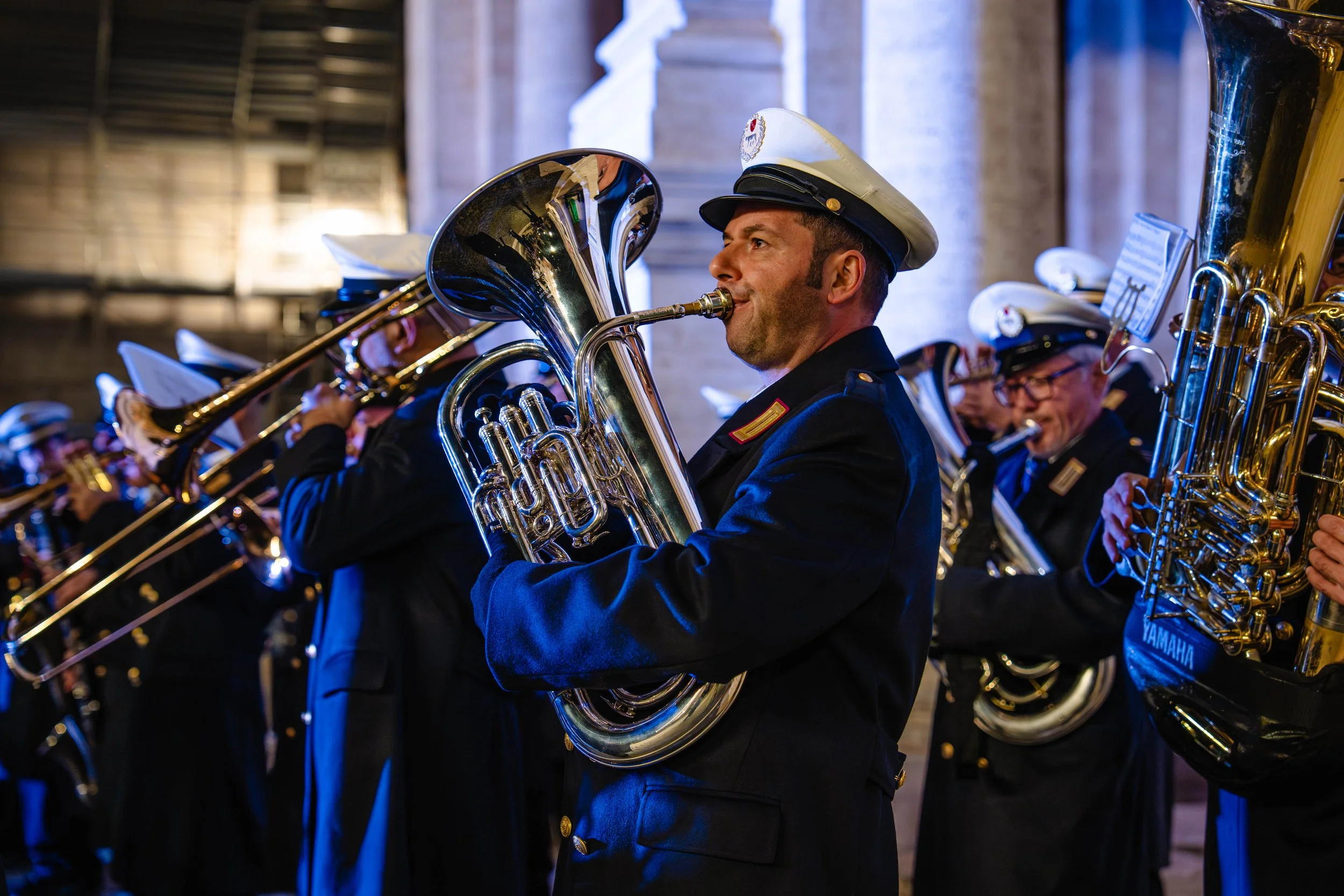 A group of musicians in uniform playing brass instruments, with the focus on a man playing a tuba, during an indoor performance.