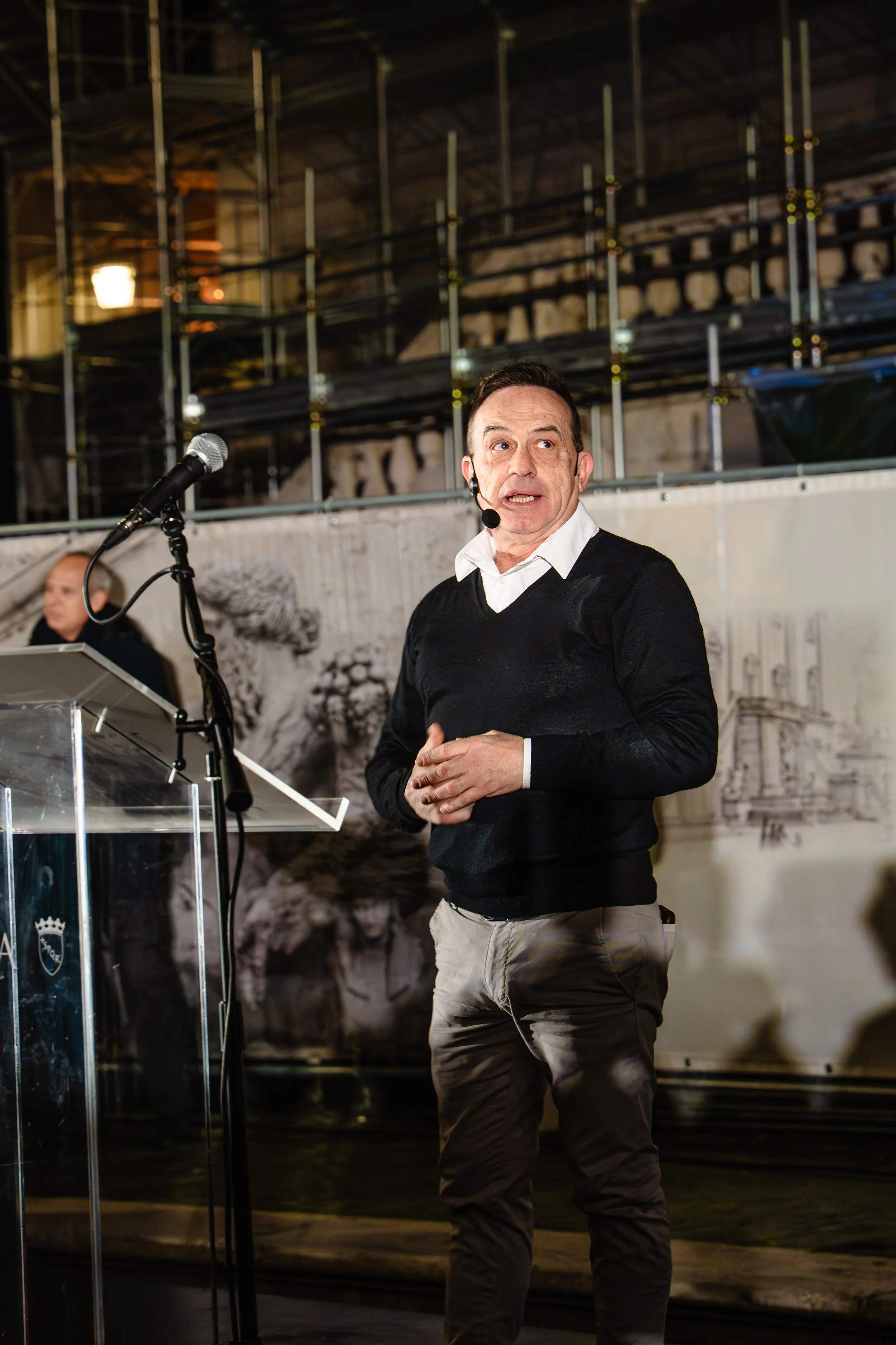 A man in a white collared shirt, black sweater, and beige pants speaking into a headset microphone at a presentation or event, with a microphone stand and a laptop nearby, in front of a backdrop with historical or architectural imagery.