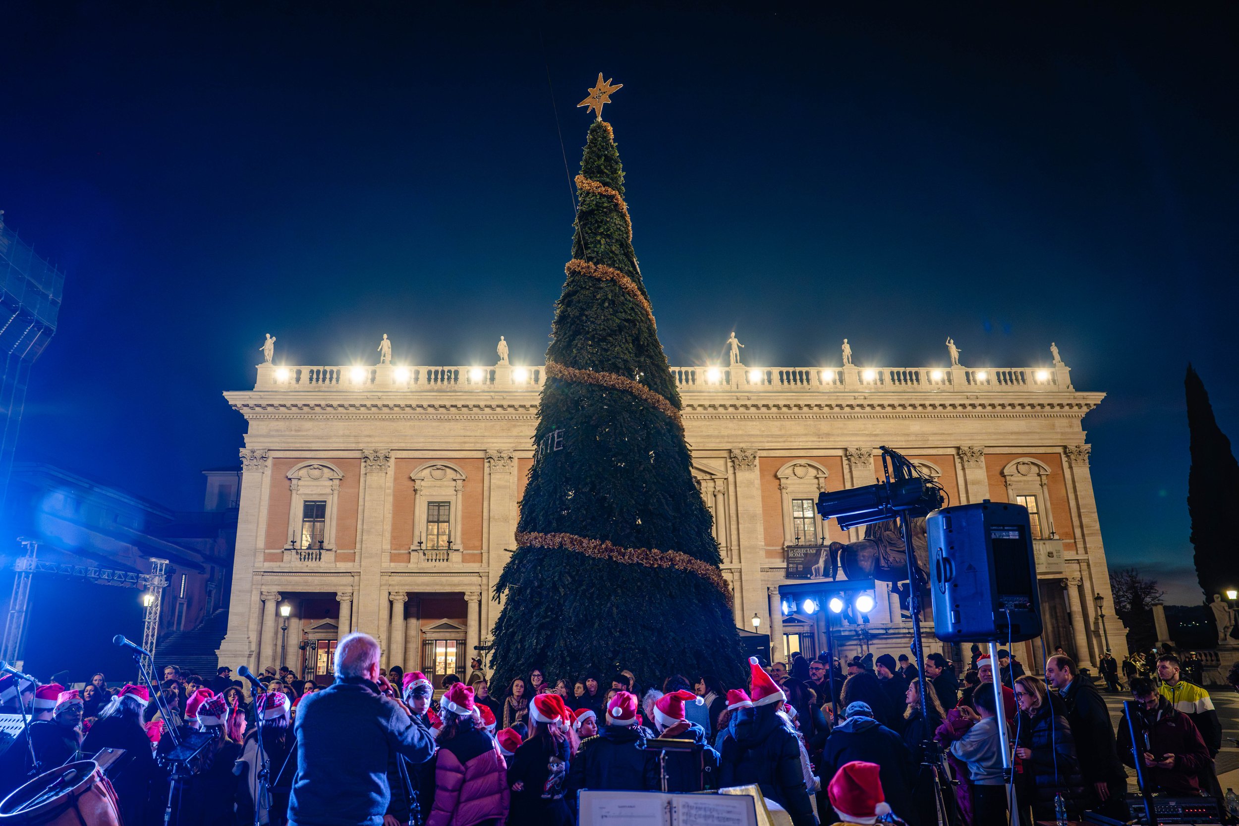 A large Christmas tree decorated with a star on top and surrounding ornaments, set up in front of a historic building with statues on the roof at night. A crowd, some wearing Santa hats, are gathered around the tree, and stage lighting illuminates th