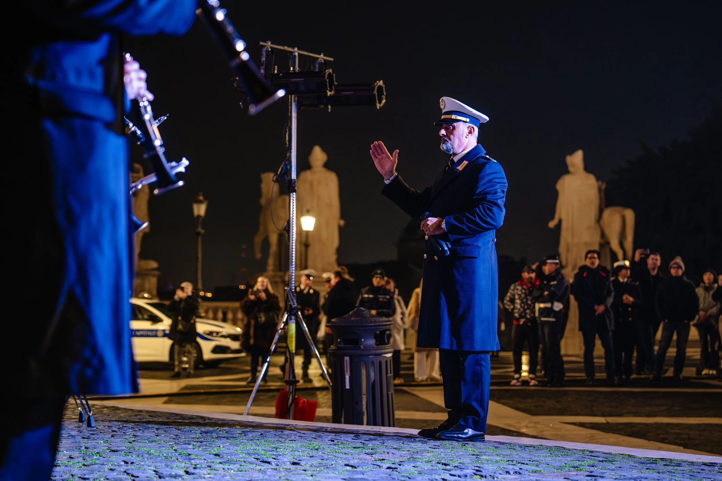 A uniformed man in a cap and coat stands with his hand raised during a night-time event with a crowd, statues, and streetlights in the background.