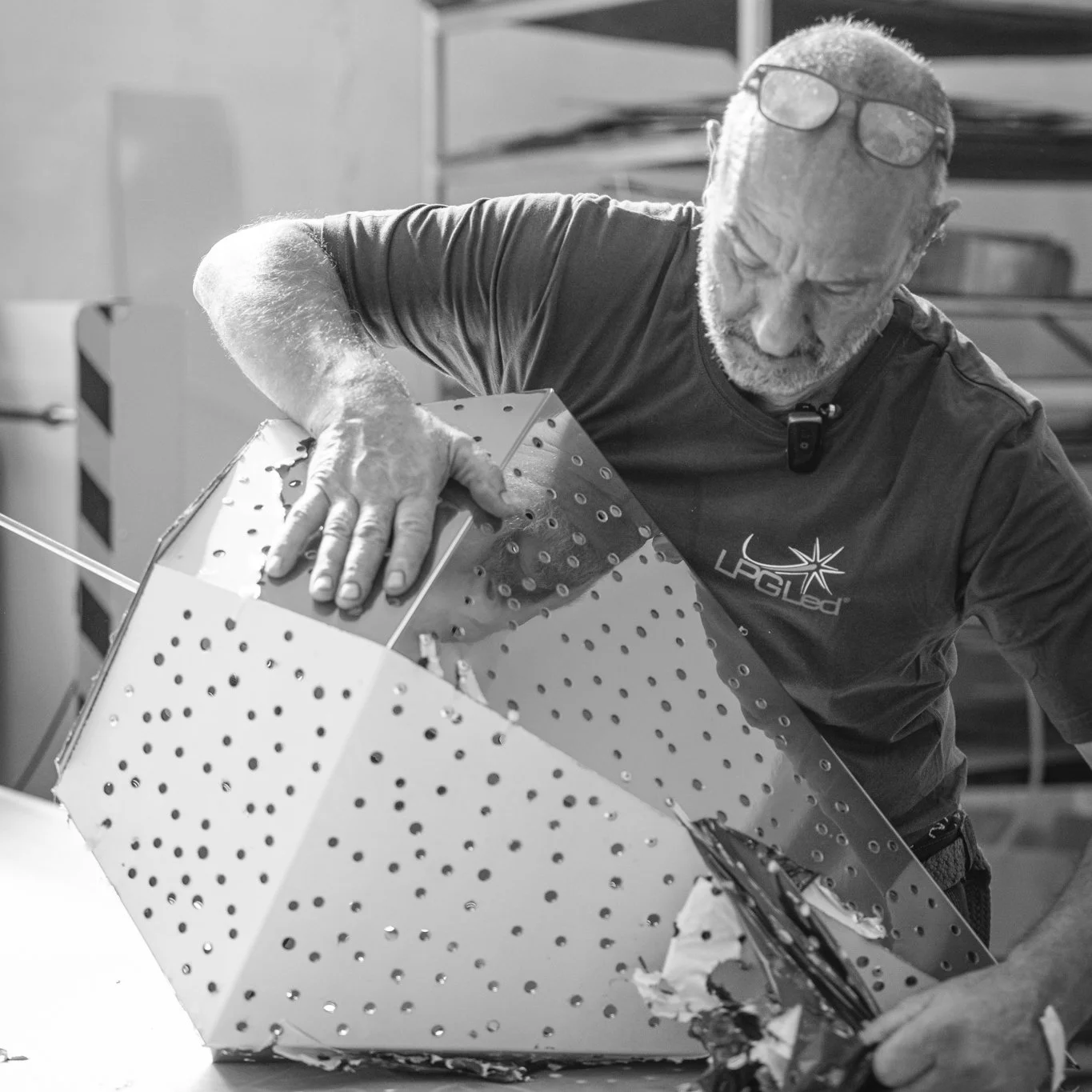 A man with glasses on his head, a beard, and a T-shirt with a logo, is working on assembling or inspecting a metal structure with perforations. He is focused on the task, with his hand placed on the metal surface. The background includes work equipme