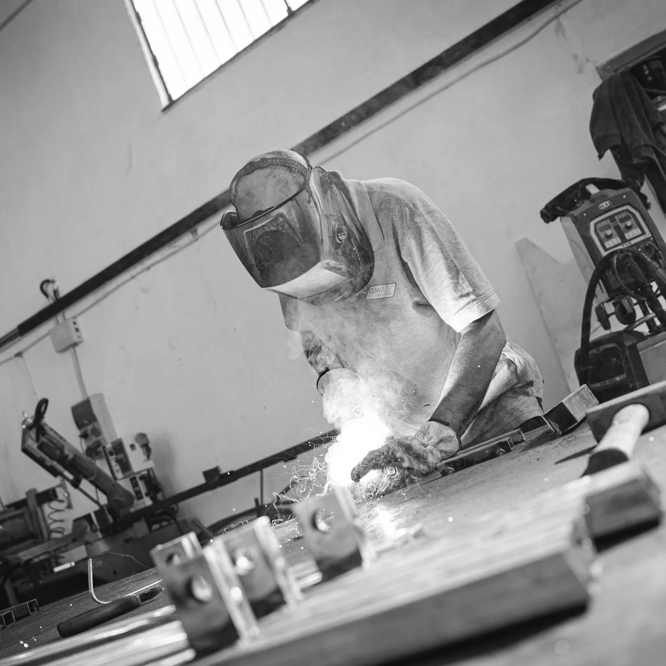 A person wearing safety goggles and a t-shirt working on a metal project in a workshop, with sparks and smoke visible.