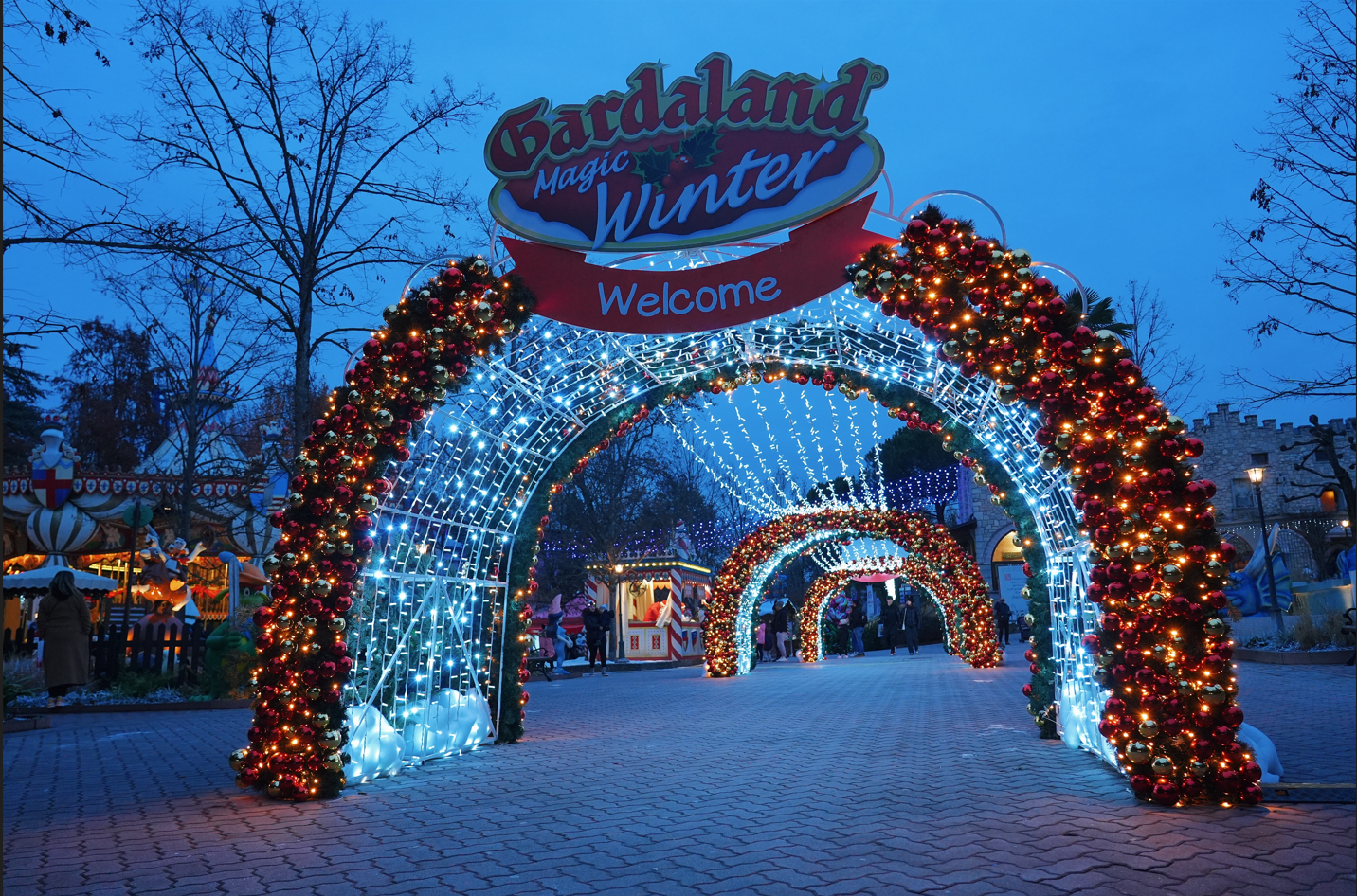 Holiday decorated park entrance with lighted arches, Christmas ornaments, and amusement rides in the background, celebrating Christmas season at Gardaland Magic Winter