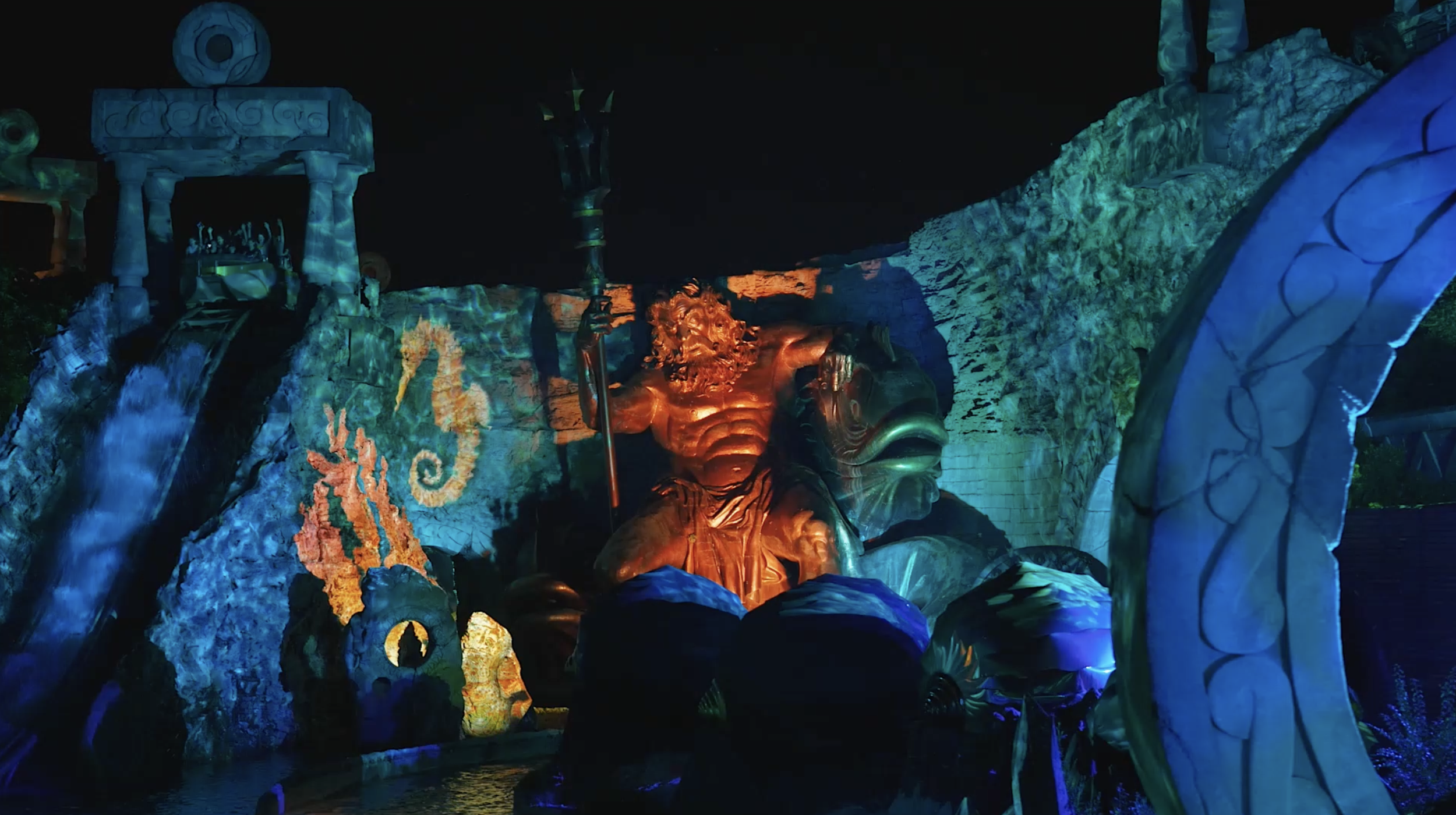Night scene of a colorful, illuminated waterfall and sculptures at a theme park, including a large lion's head and tropical plants.