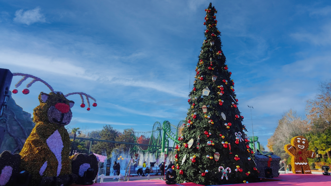A decorated Christmas tree in an outdoor amusement park with holiday figures, including a reindeer and gingerbread person, and a roller coaster in the background under a blue sky.