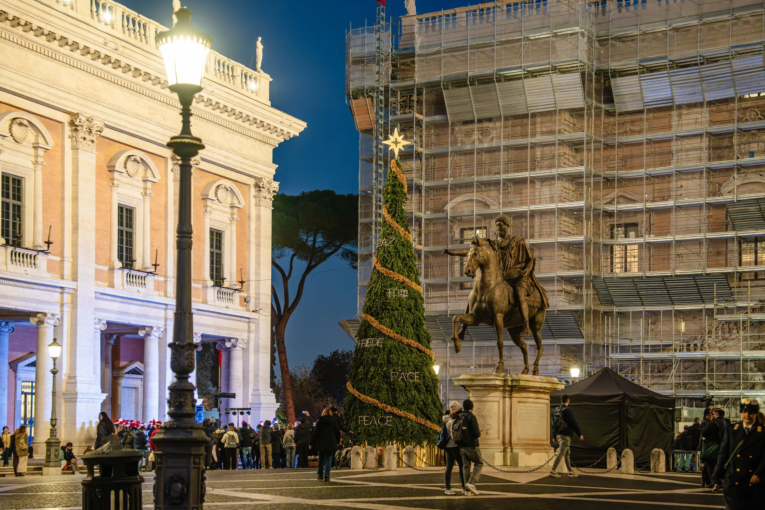 A large Christmas tree decorated with garlands and a star on top stands in front of a historic building under renovation, with scaffolding. People gather in the public square, some wearing festive clothing, during evening hours.