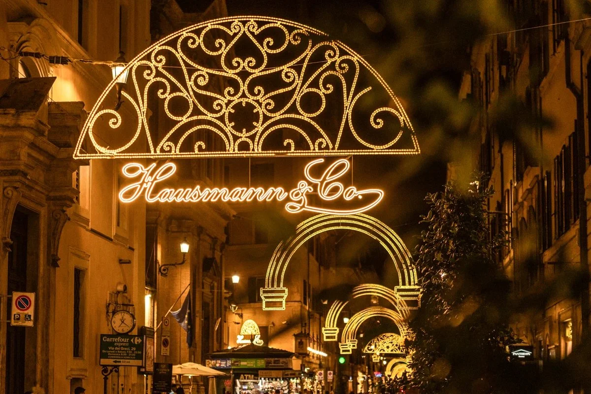 Illuminated decorative sign with intricate gold swirling patterns hanging over a street at night, with the words 'Hausmann & Co.' written at the bottom.