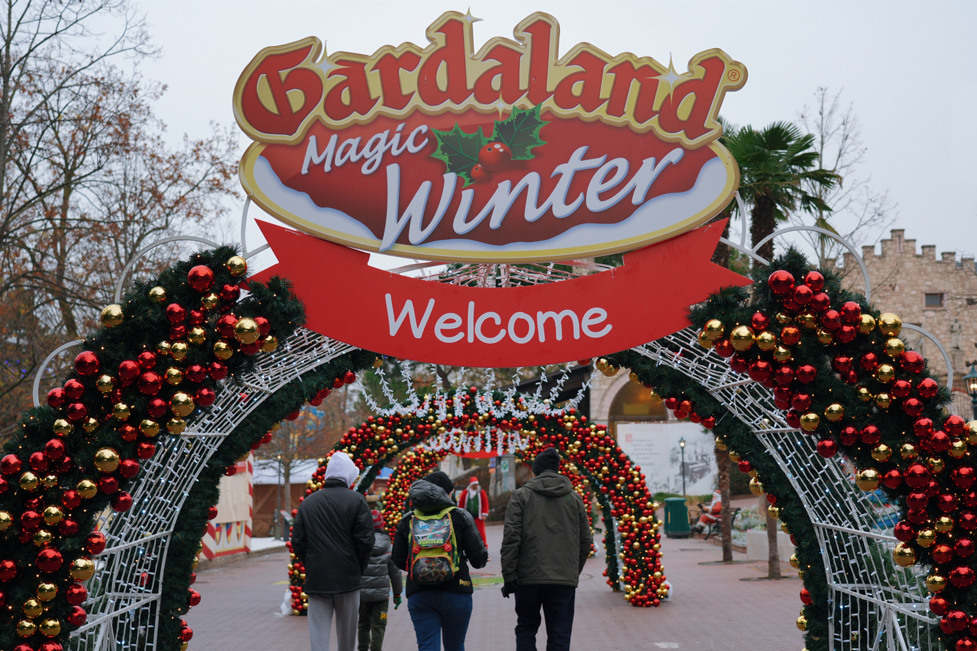 Entrance arch decorated with red and gold Christmas ornaments at Gardaland Magic Winter, with visitors walking underneath, Christmas trees and rides visible in the background.