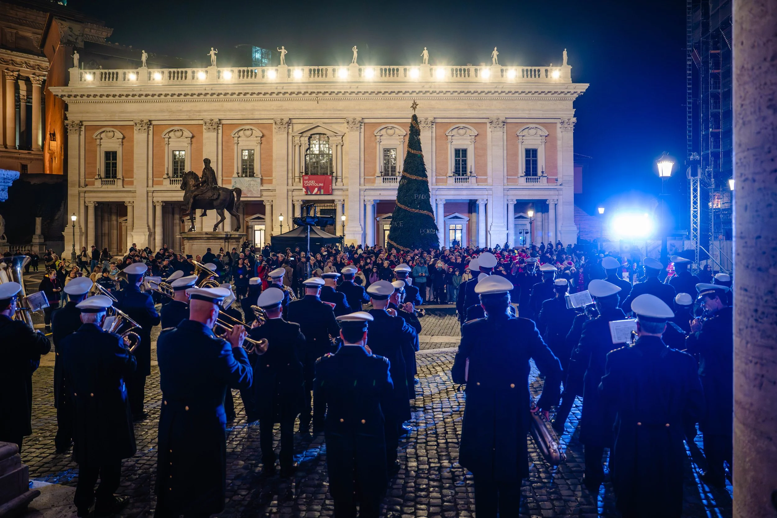 Nighttime scene of a large crowd gathered in front of a historic building with classical architecture, a Christmas tree, and a statue of a horse and rider. The scene includes a marching band in uniform, bright lights, and festive decorations.