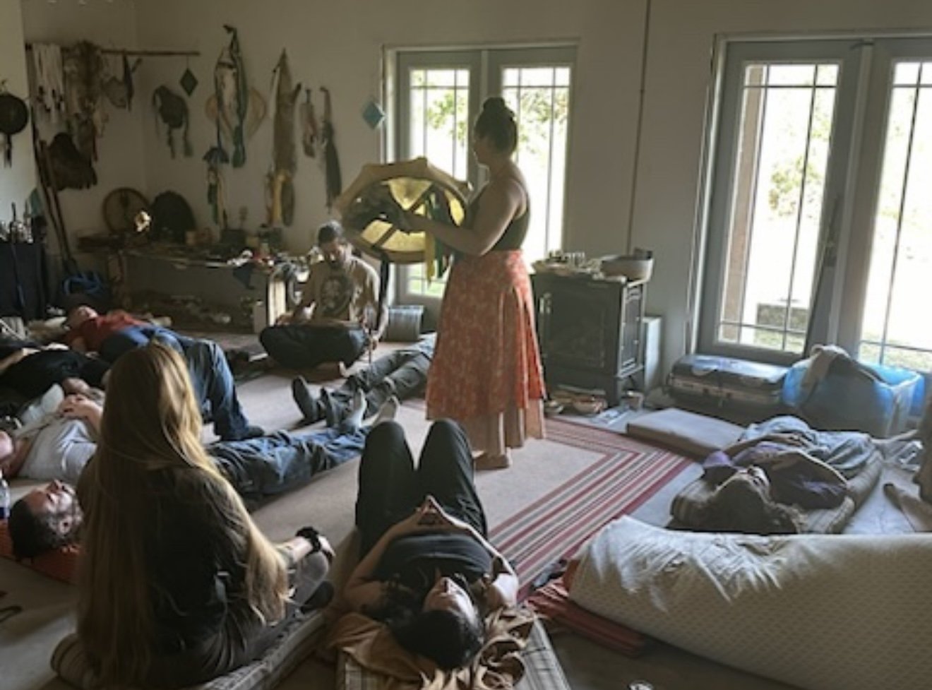 Group of young people lying and sitting on mats and blankets in a cozy, sunlit room, engaging in a meditation or mindfulness session, with a woman in a long skirt leading the activity, surrounded by wall decorations, a television, and large windows s