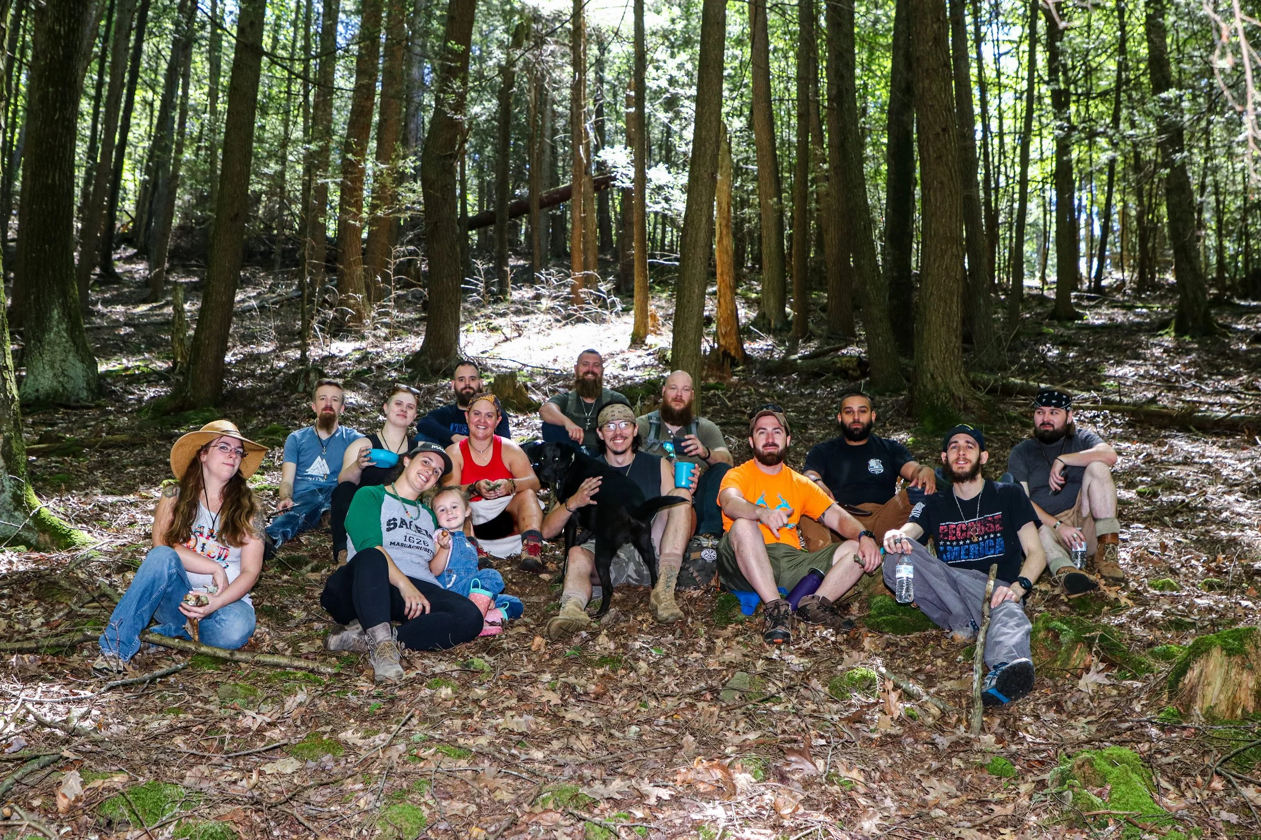 Group of people sitting on forest ground, smiling, with trees and sunlight in the background