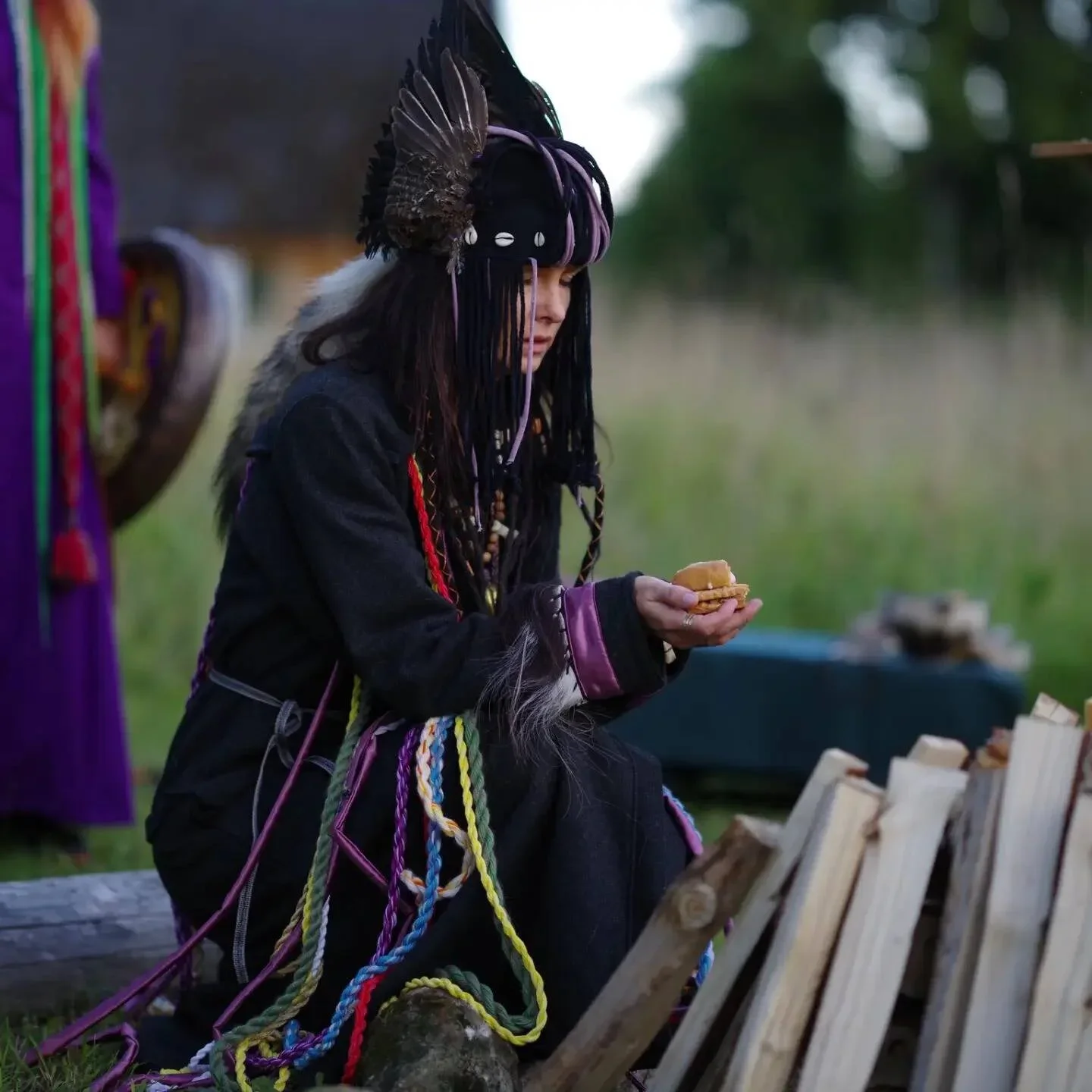 A person wearing traditional indigenous attire, including a feathered headdress and beaded decorations, is sitting outdoors near a stack of cut wood, holding a small object or food in their hand.