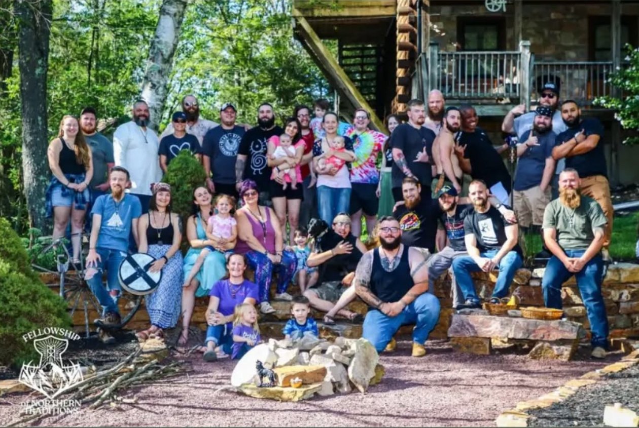 Large group of people outdoors on a sunny day, posing for a photo in front of a wooden building, with trees and greenery in the background.