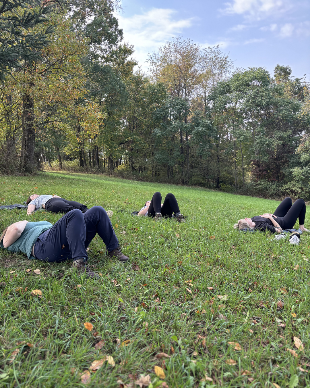 Four people lying on a grassy field during a yoga or meditation session, surrounded by trees and under a cloudy sky.