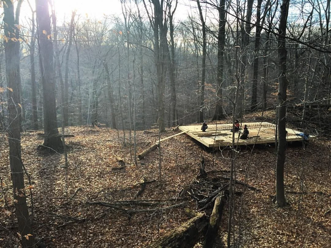 A group of people working on constructing a wooden platform in a wooded forest area during late autumn, with fallen leaves on the ground and leafless trees surrounding them. Perfect places for shamanic, animistic, yogic, and wellness retreats.