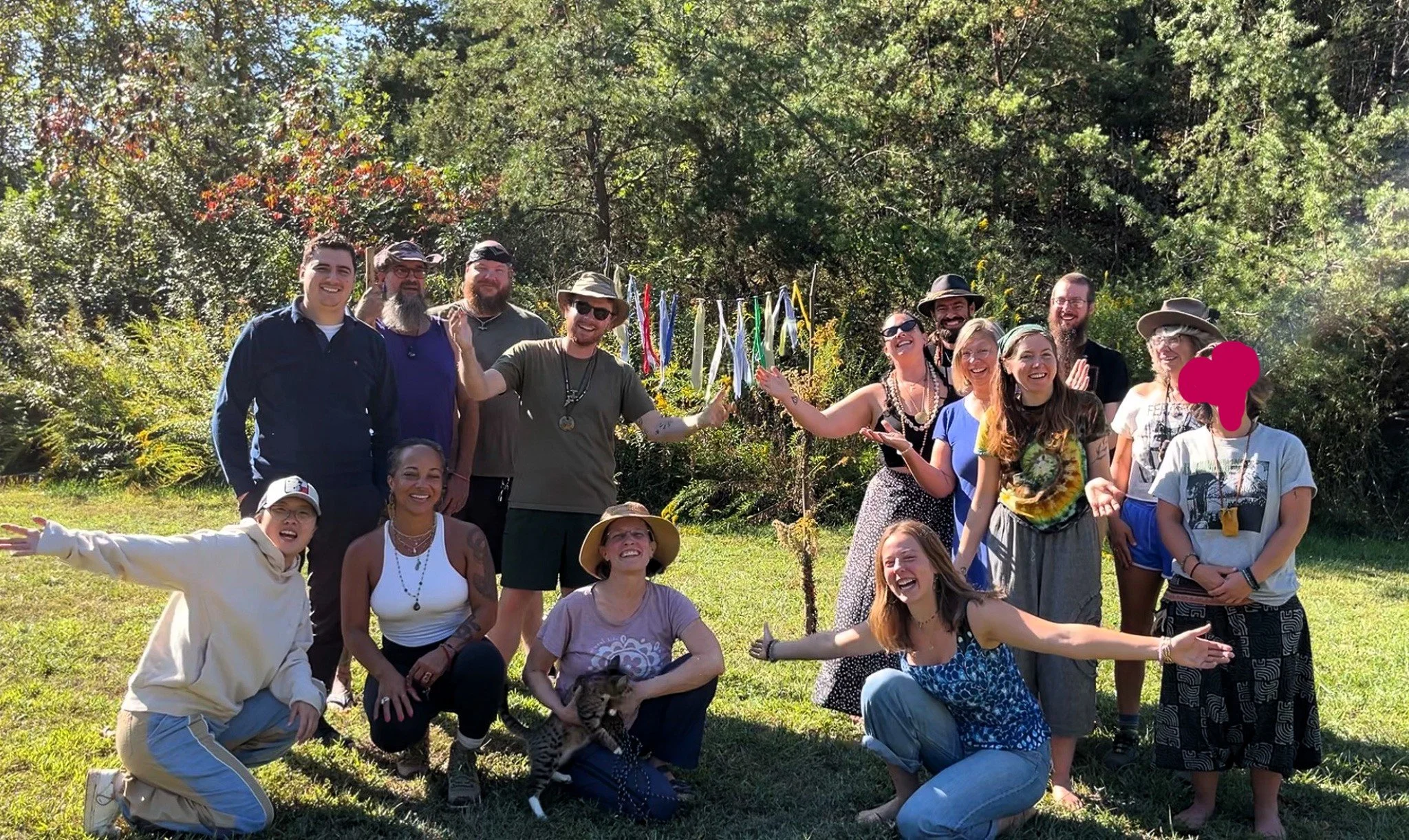 A group of 15 diverse people outdoors, smiling and celebrating, with colorful ribbons on a tree in the background.
