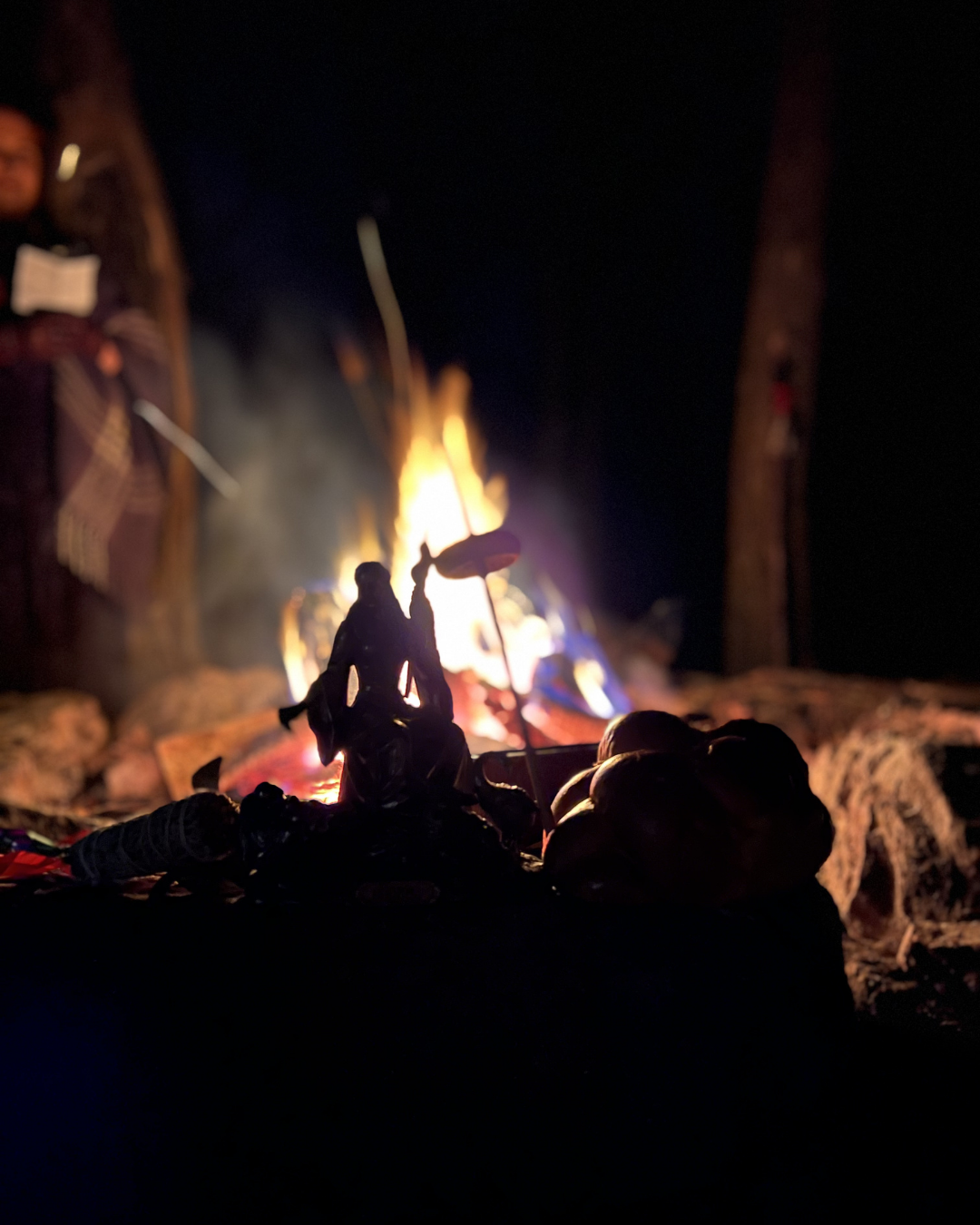 Silhouette of a figurine sitting on a surface with a campfire in the background at night.