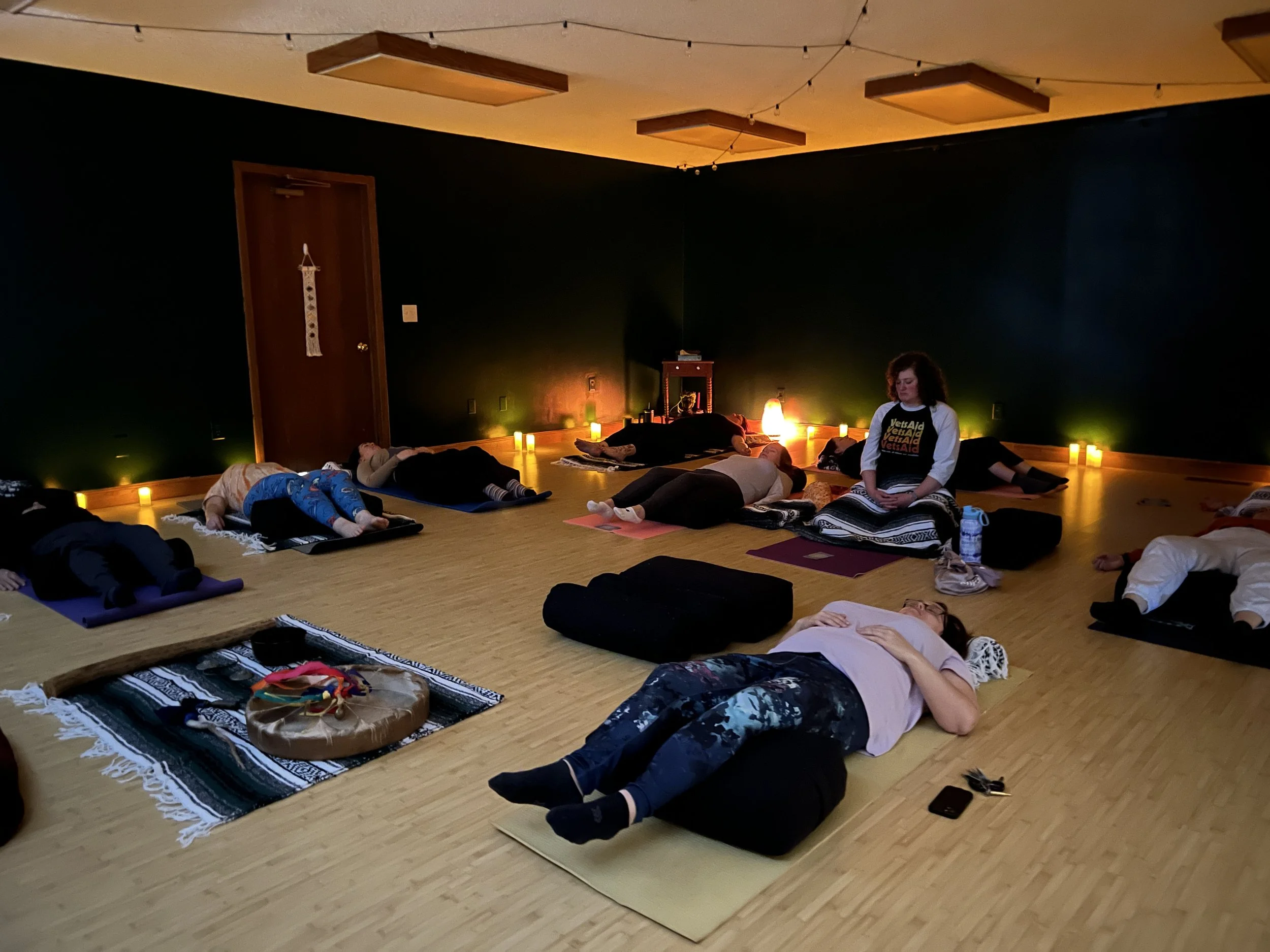 People practicing yoga in a dimly lit studio with candles, mats, and bolsters, with a woman in center sitting cross-legged.