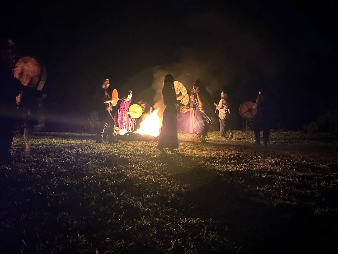 People gathered around a bonfire at night, some holding drums and others standing, with smoke and darkness surrounding them.