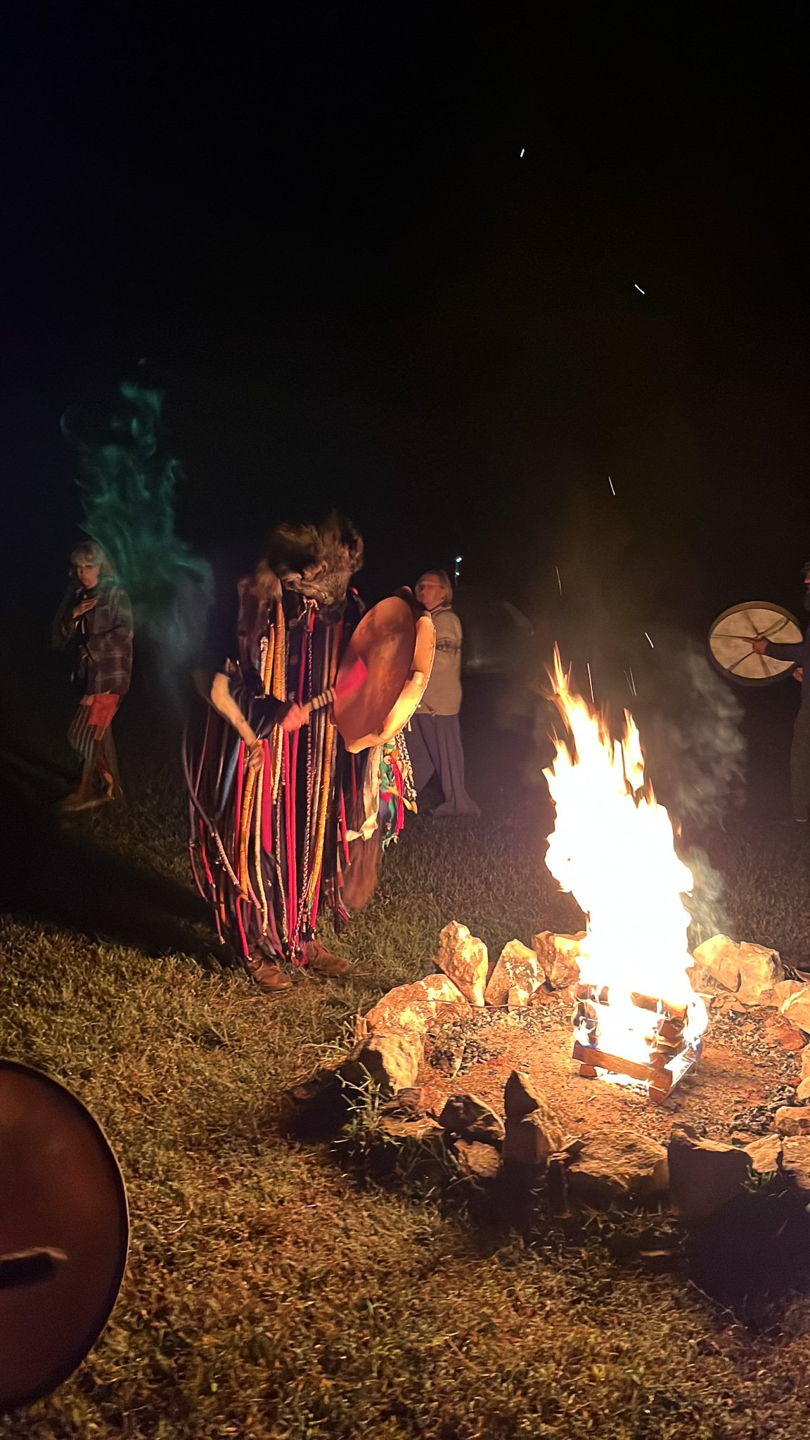 People participating in a Native American ceremony around a campfire at night, with traditional clothing and musical instruments.