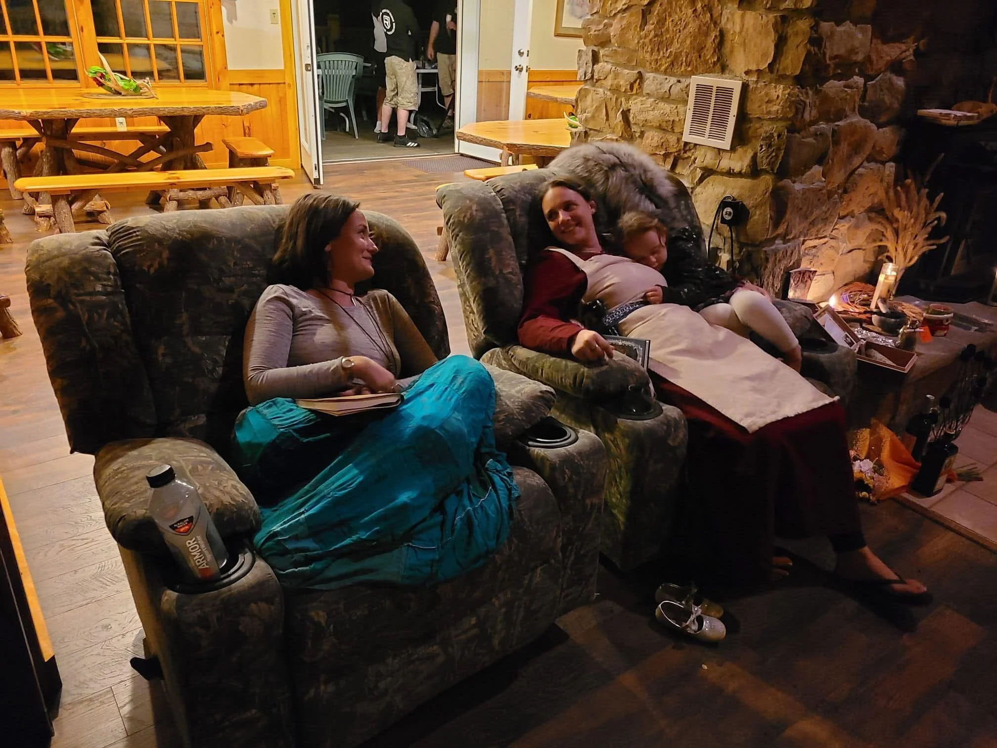 Three people relaxing on sofas in a cozy room, with a stone fireplace and a table with decorations nearby. Two women and a child are visible, one woman with a blue skirt sitting on the left, another woman with a white apron sitting in the middle, and