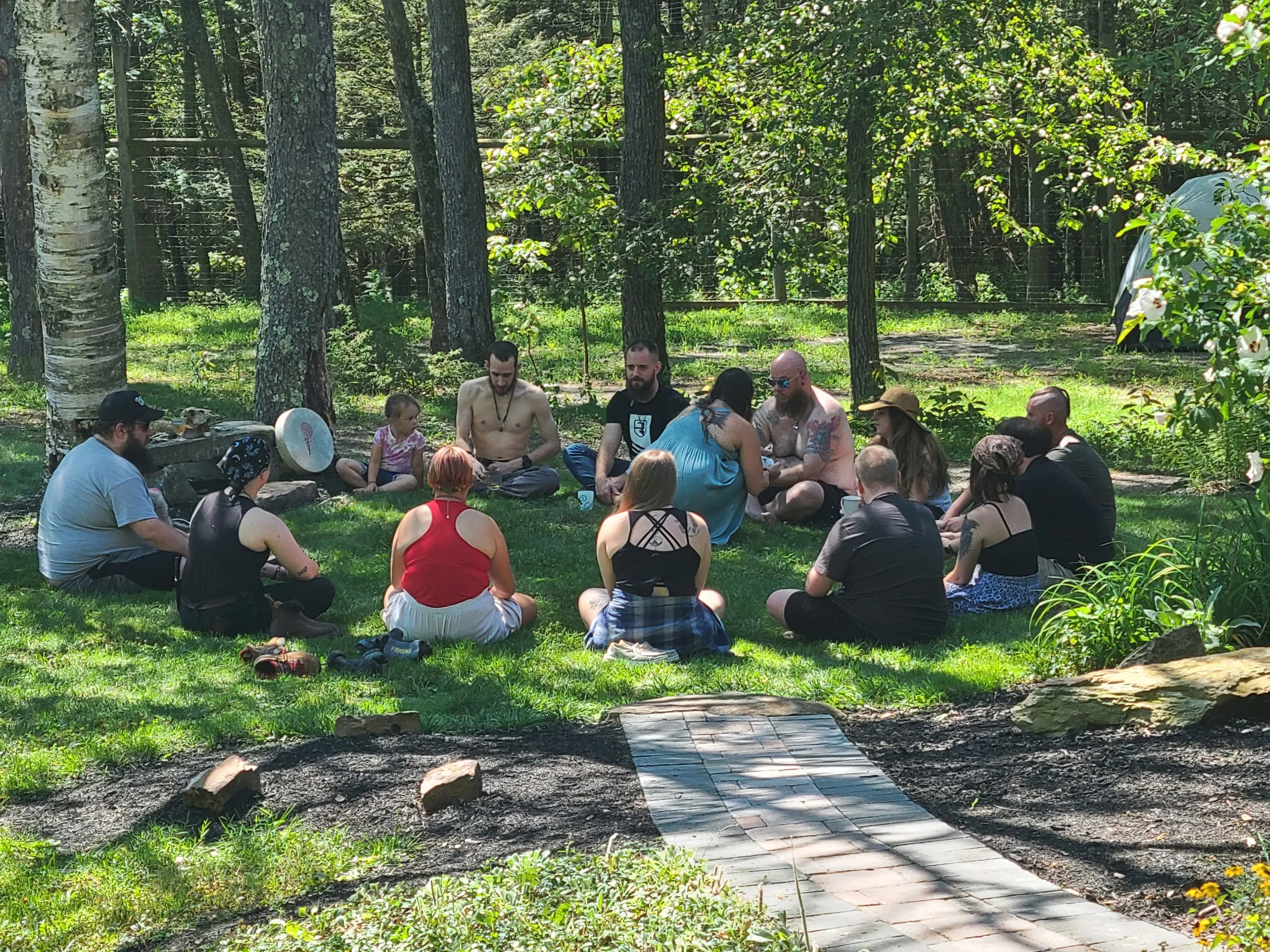 A group of people sitting in a circle outdoors in a forested area on grass, participating in a communal activity.