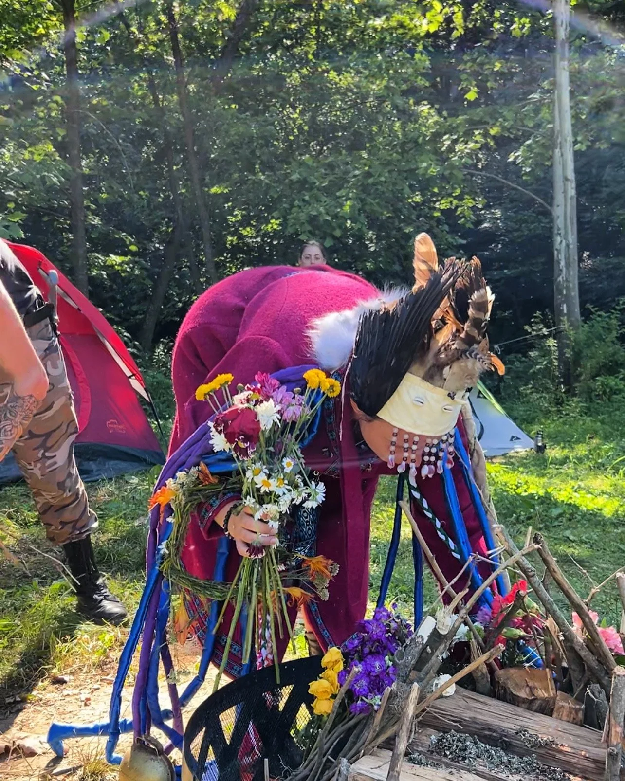 Person wearing a tribal headdress and colorful clothing placing flowers on a pile of wood at an outdoor site with tents and green trees in the background.