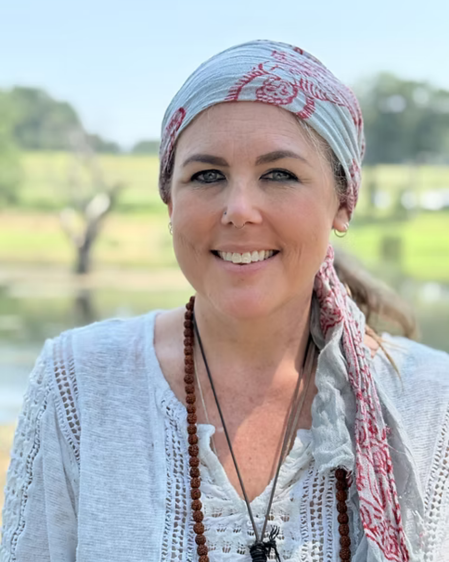 A woman with light skin, blue eyes, and a nose piercing smiling outdoors, wearing a light-colored headscarf with red patterns and a white embroidered top, with a blurred green landscape in the background.
