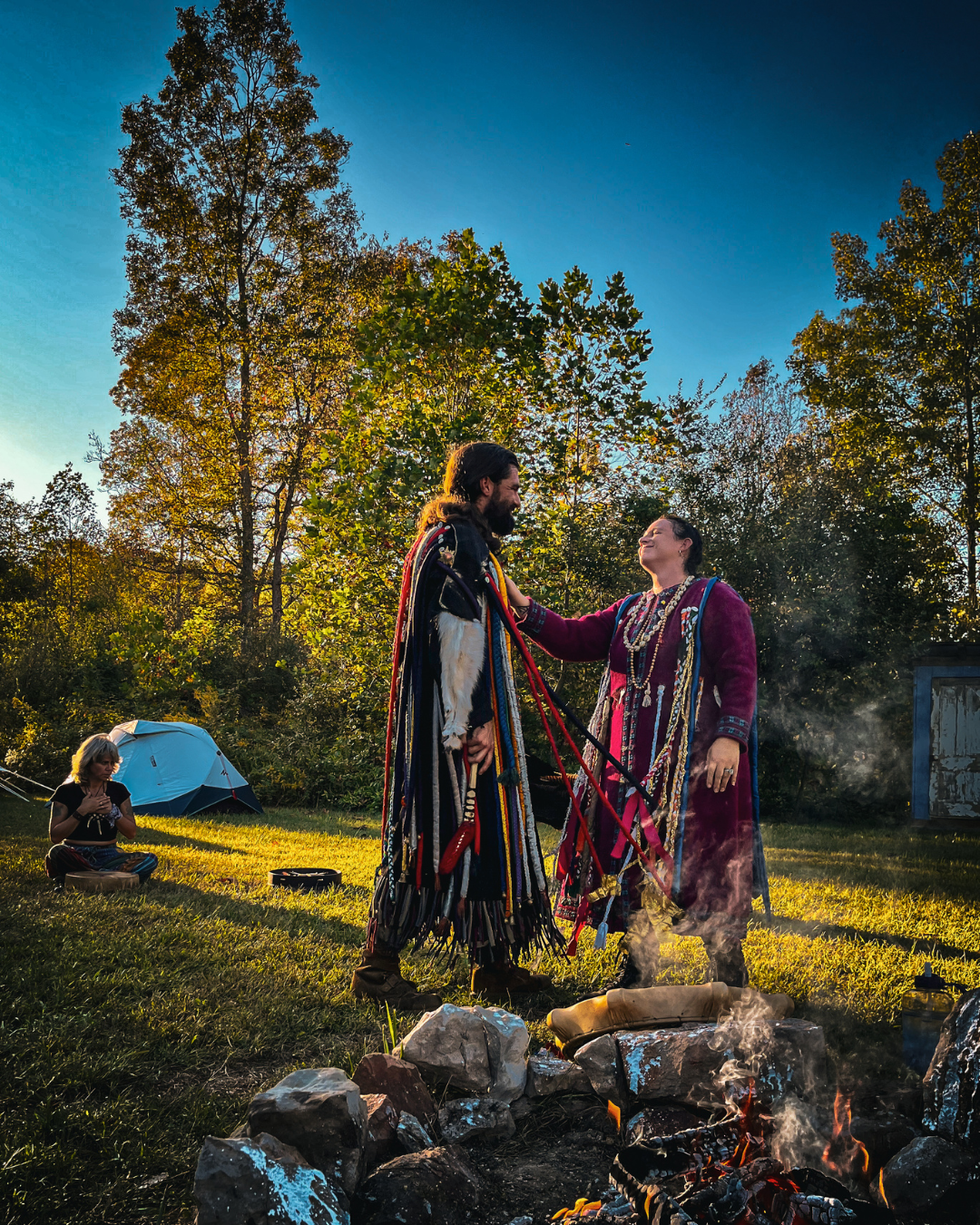 Two people in traditional Native American attire standing by a campfire, with a woman sitting on the grass near an umbrella in the background, surrounded by trees during sunset.