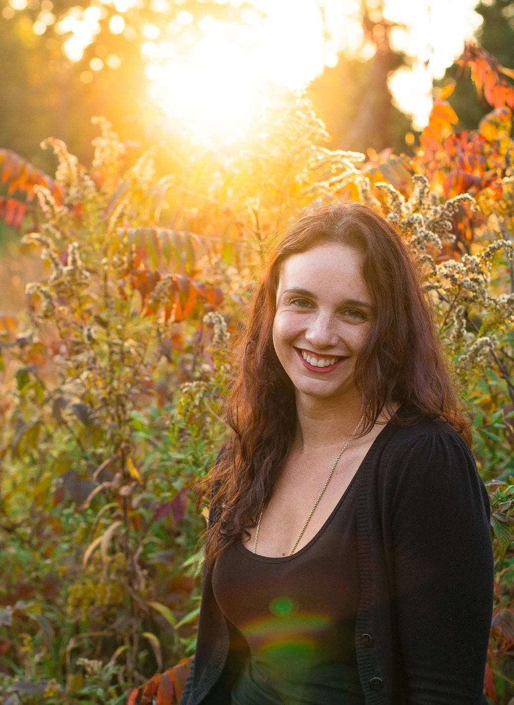 A young woman with long curly hair smiling outdoors during sunset, standing in front of autumnal foliage.