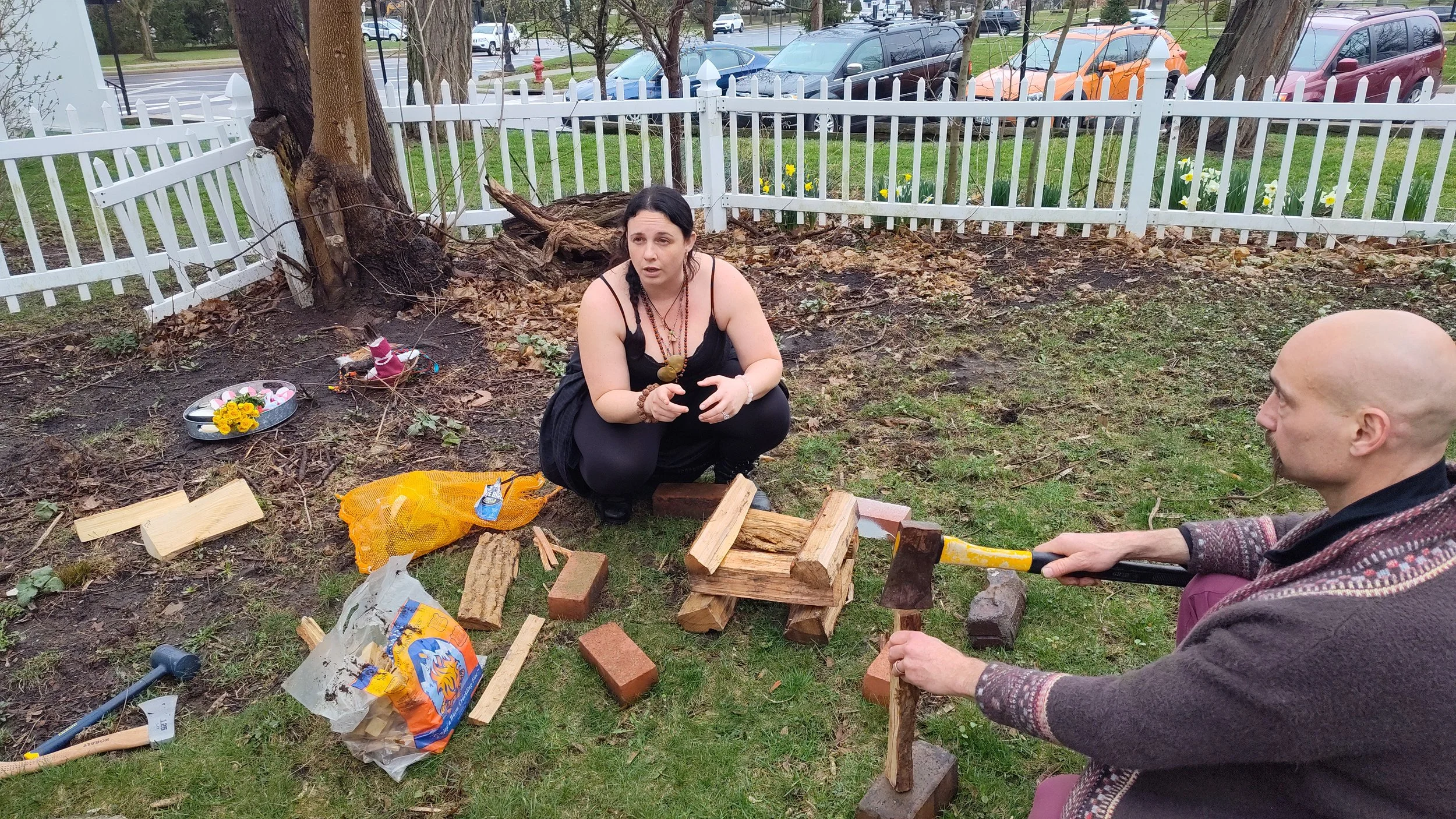 A woman squatting in a yard with a white fence, surrounded by logs and bricks, talking to a bald man who is holding an axe raised above a log. There are tools, a bag of wood, and gardening tools scattered around.