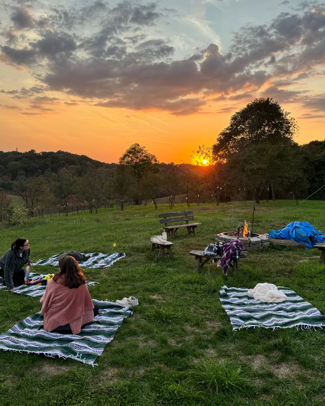 Two people sitting on blankets around a fire pit at sunset in a grassy open area with trees and distant hills.