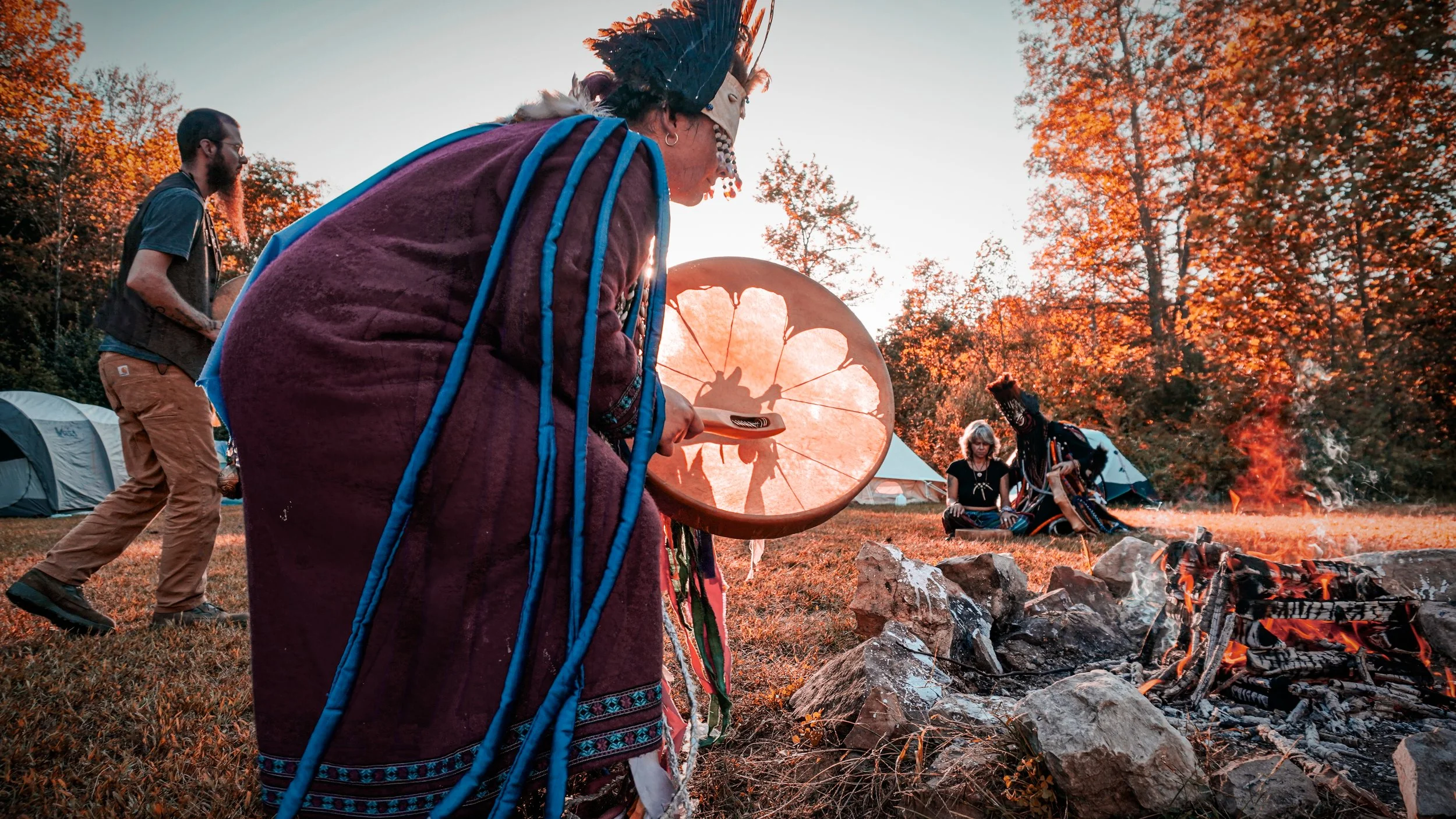 Indigenous people performing a ceremonial dance around a campfire in a forested area during sunset.