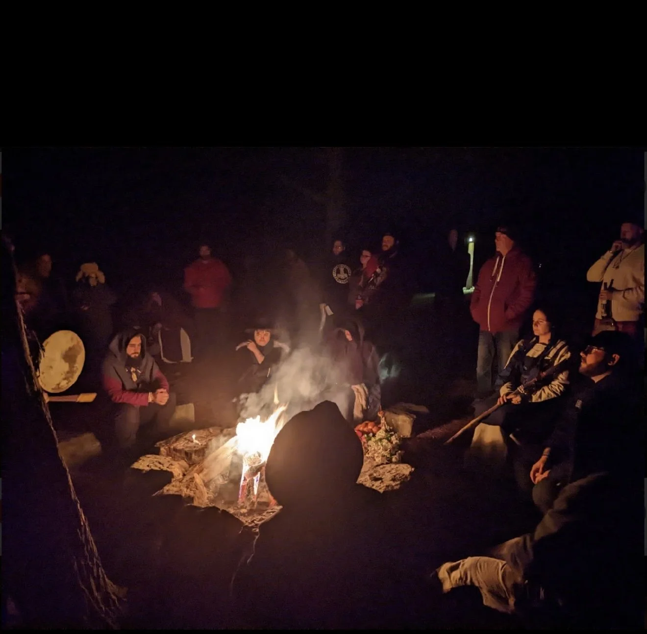 Group of people gathered around a campfire at night, some sitting and some standing, outdoors in a dark setting.