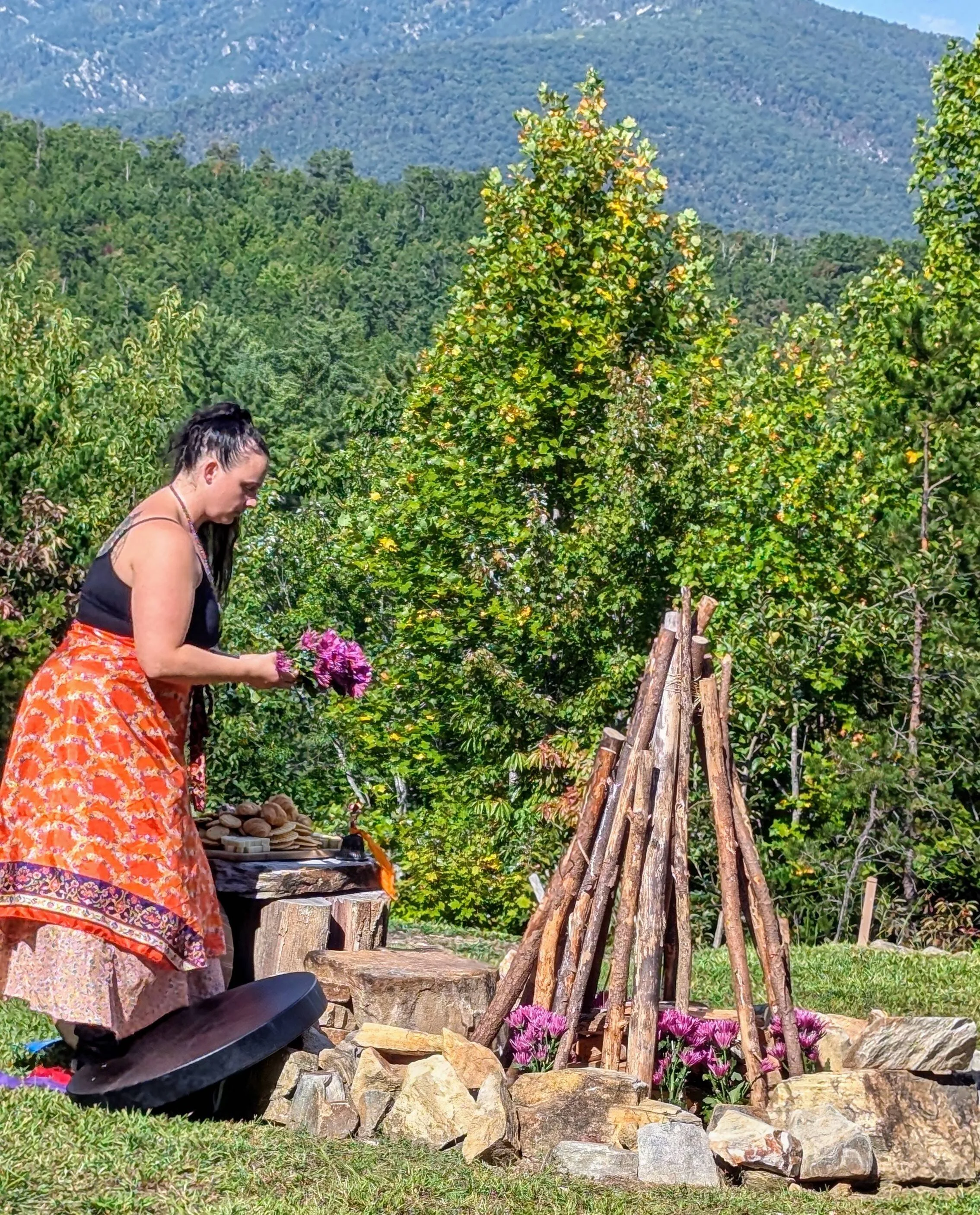 A woman dressed in a colorful skirt and black top is standing outdoors on a grassy area, arranging flowers near a wooden teepee-like structure made of logs. The scene is set in a lush green environment with trees, bushes, and mountains in the background.