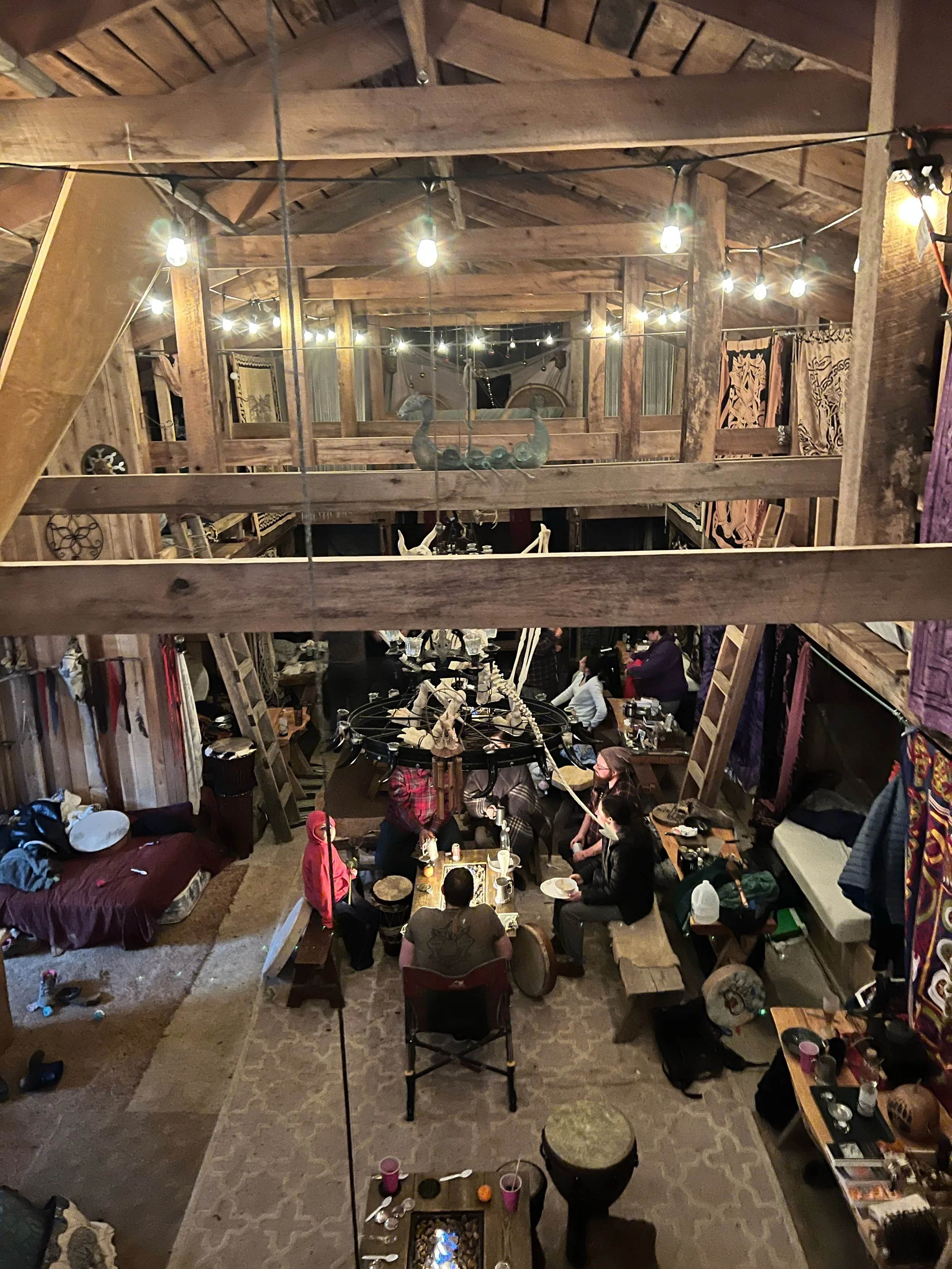 People gathered around a dining table with food and drinks in a rustic, wood-paneled loft with string lights hanging from the ceiling.