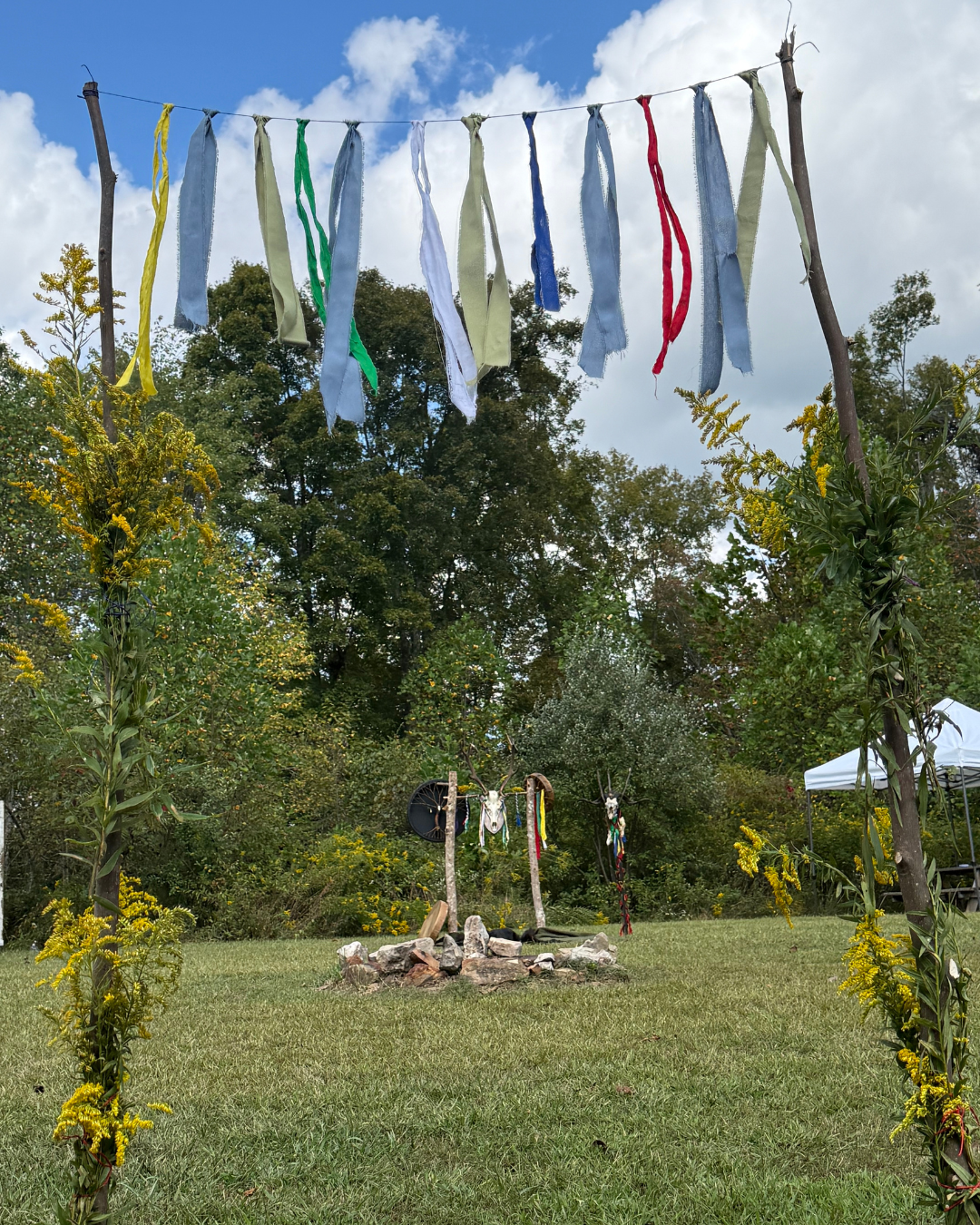 An outdoor scene with fabric strips hanging from a rope between two wooden posts, a small stone fire pit with decorated sticks around it, yellow flowers, trees, and a white tent in the background.