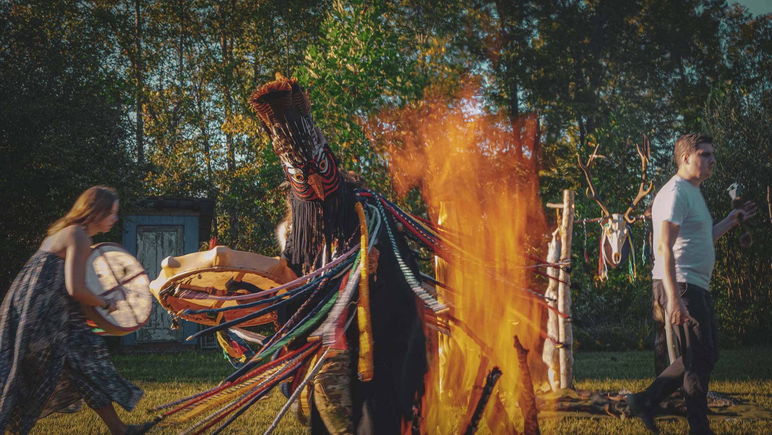People participating in a traditional shamanic ritual with Ayan Kam outdoors, with a large bonfire in the center and decorated masks hanging on a wooden structure in the background.