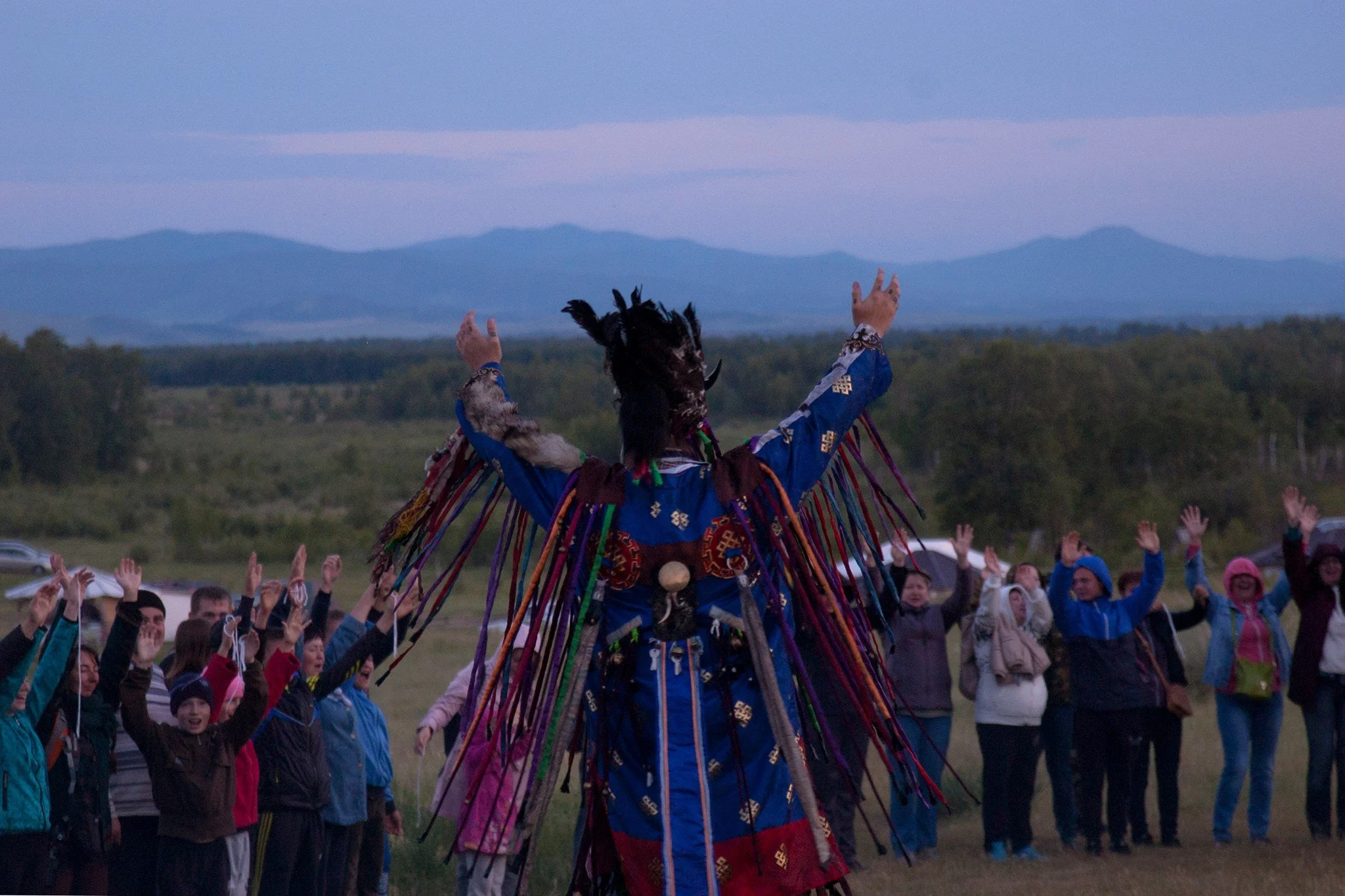 A person dressed in traditional Indigenous attire with fringes and decorations leading a crowd in an outdoor ceremony during dusk, with mountains in the background.