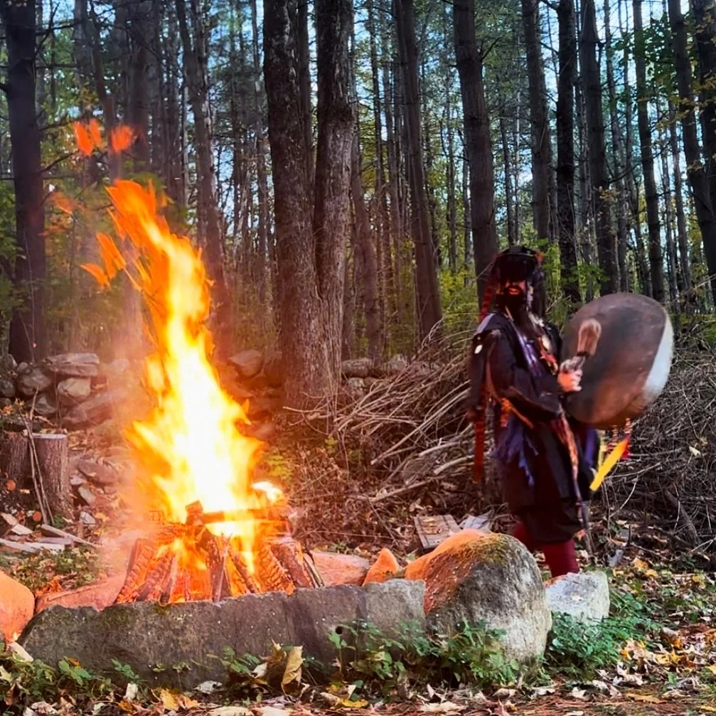 A person dressed in costume standing next to a large campfire in a forest set. The person holds a shield and a weapon, wearing a hat with horns and long hair, resembling a Viking or medieval warrior.