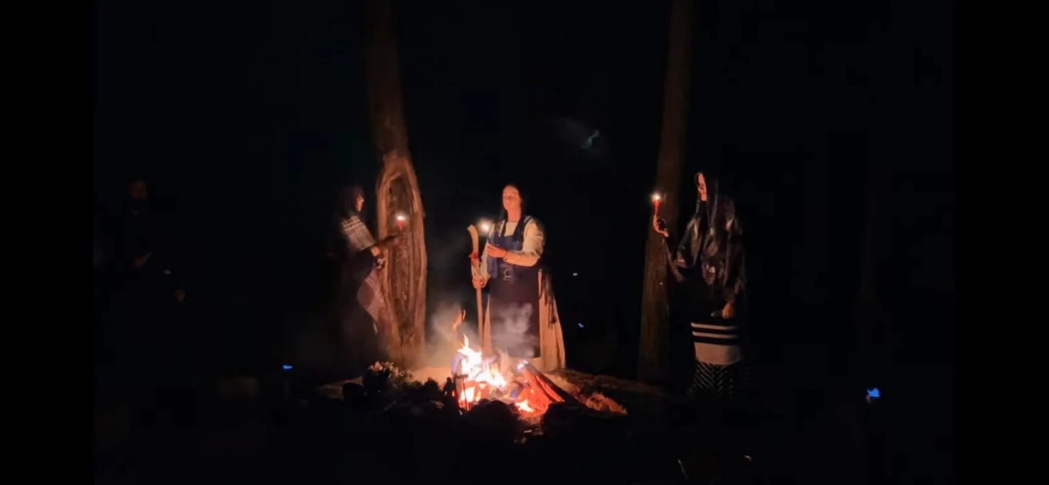 Three women standing around a small campfire at night, holding candles, with two large trees in the background.