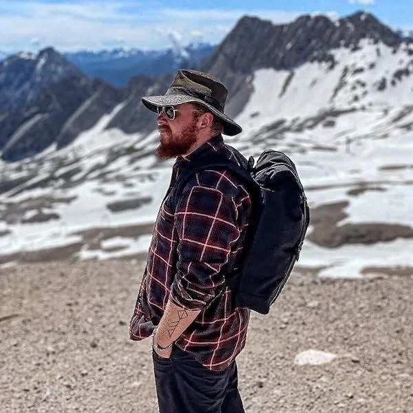 A man with a beard wearing a hat, sunglasses, a plaid shirt, and a backpack, standing outdoors in a snowy mountainous landscape.