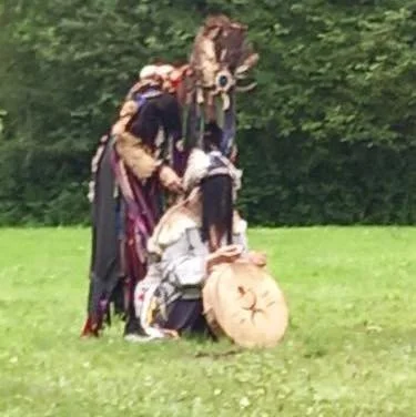 Two people wearing historical costumes and a person in modern clothes gathered outdoors with a large wooden drum.