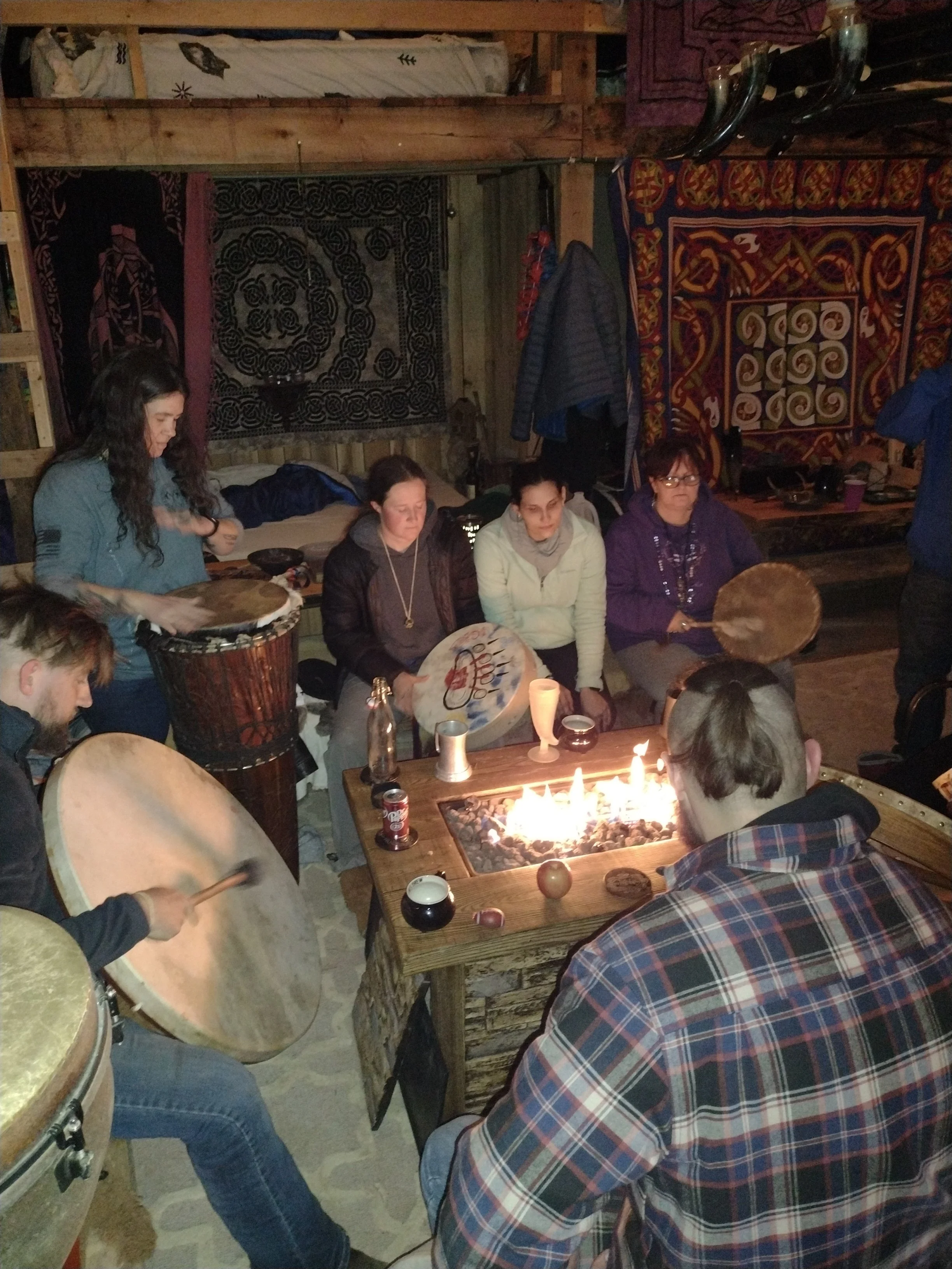 Group of people sitting around a fire pit inside a rustic wooden room, with some playing drums and others sitting and watching.