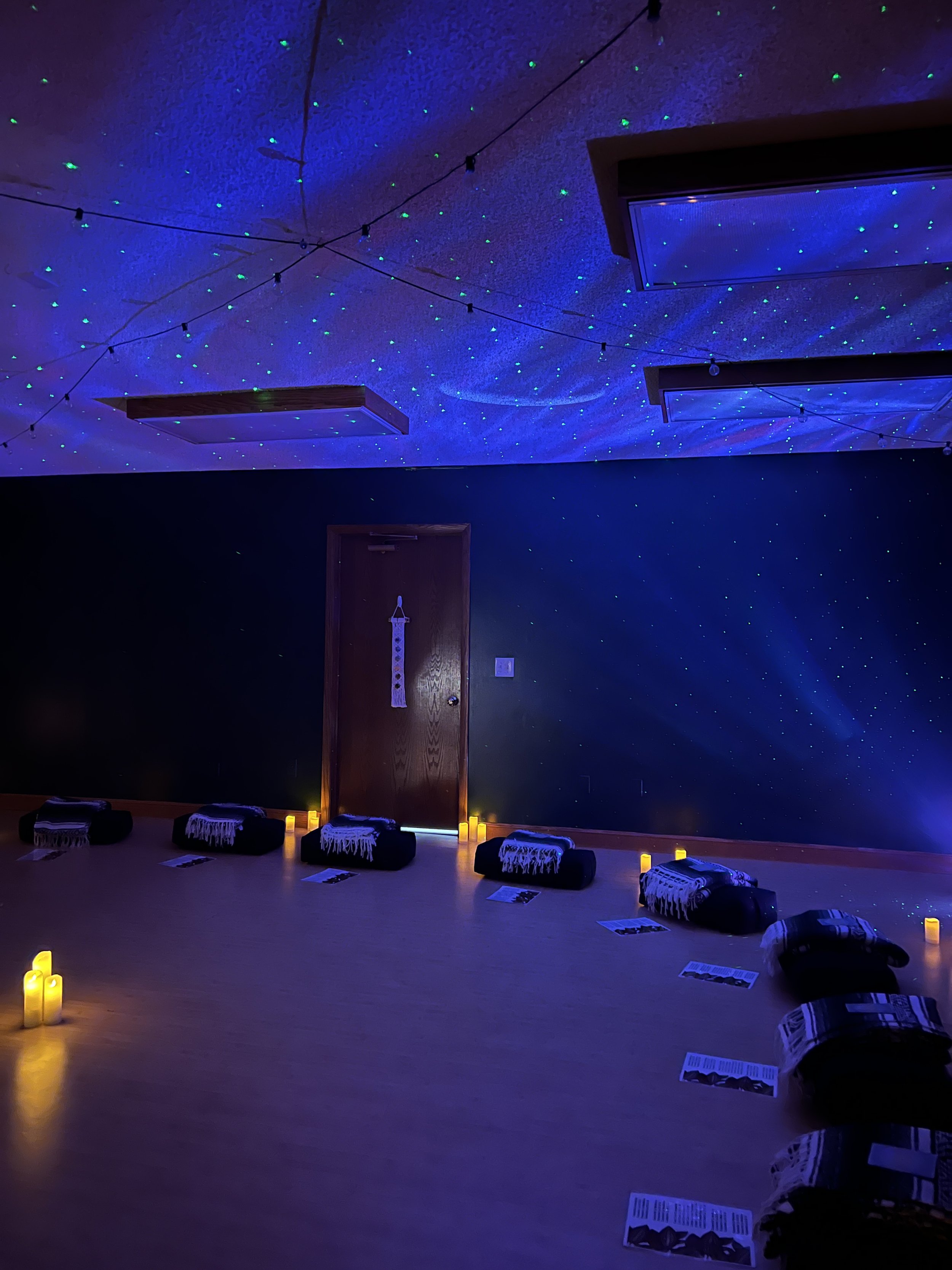 A dimly lit room with glowing candles along the floor in a semicircle, and fabric items with informational pamphlets in front of each. The ceiling is decorated with tiny blue and green lights resembling stars, with string lights hanging across the ro