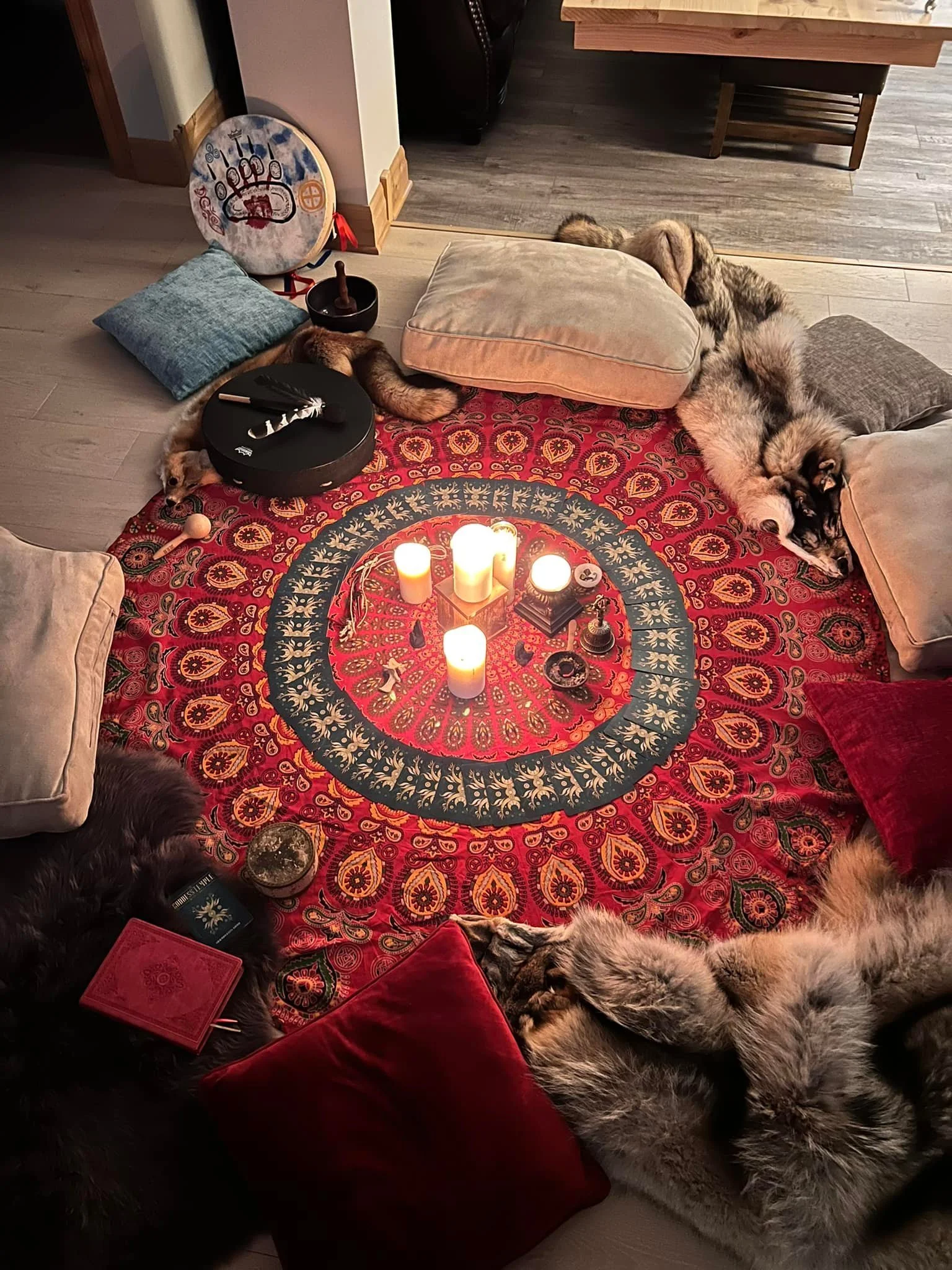 A cozy, dimly lit space with a red patterned round blanket on the floor, candles in the center, surrounded by several pillows and blankets, and multiple dogs resting around the setup.
