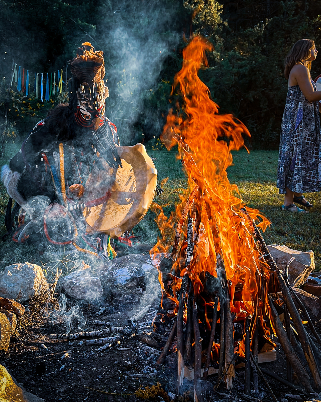 A person in traditional Native American attire playing a drum near a large bonfire outdoors at dusk, with a woman in a patterned dress standing nearby.