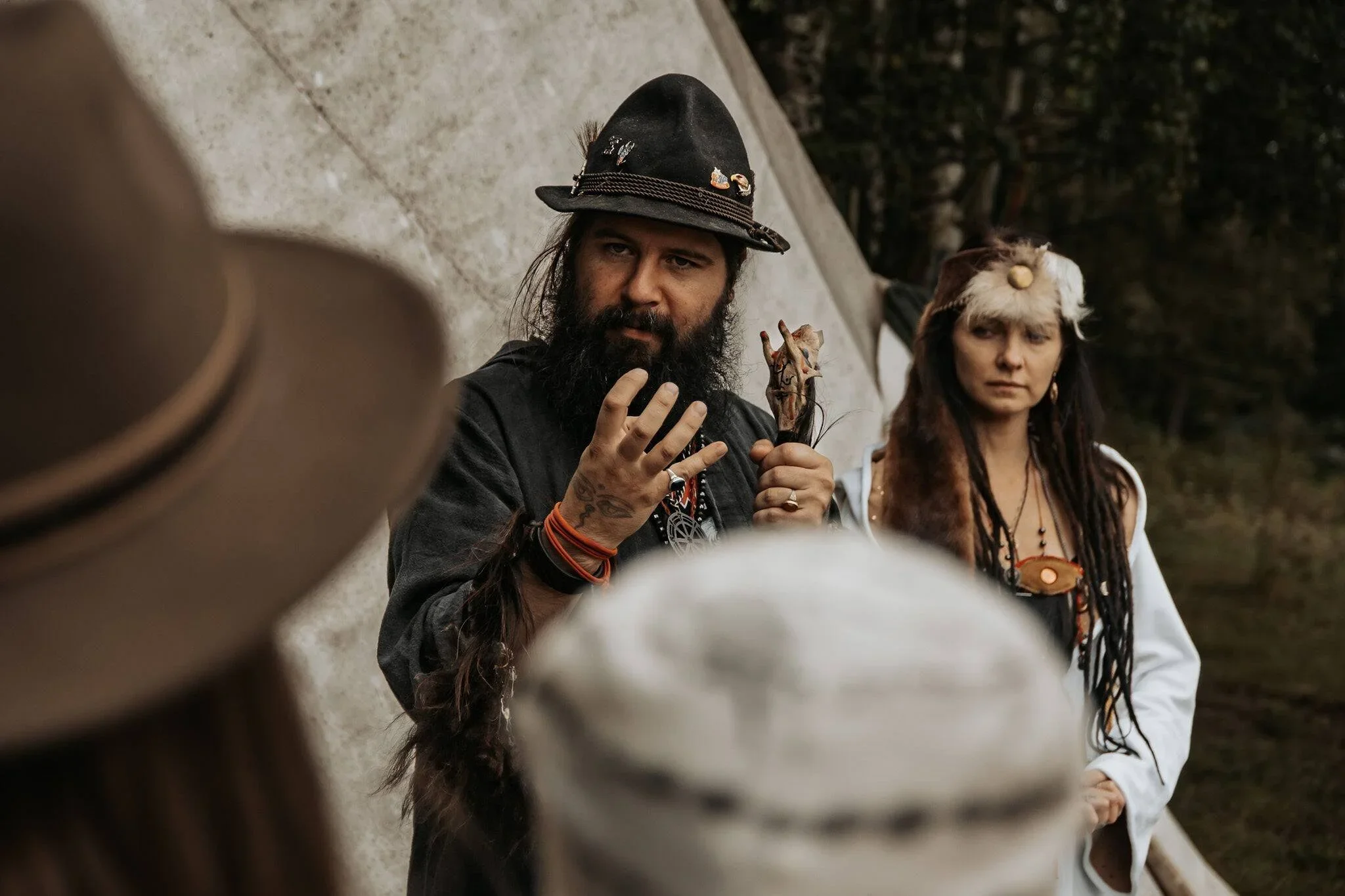 A man with a long beard, black hat decorated with pins, and dark clothing holding a small puppet or figurine, standing near a woman with long hair and dreadlocks, wearing a white jacket and a headband with a feather. They are outdoors, partially obscured by a person in the foreground wearing a hat.